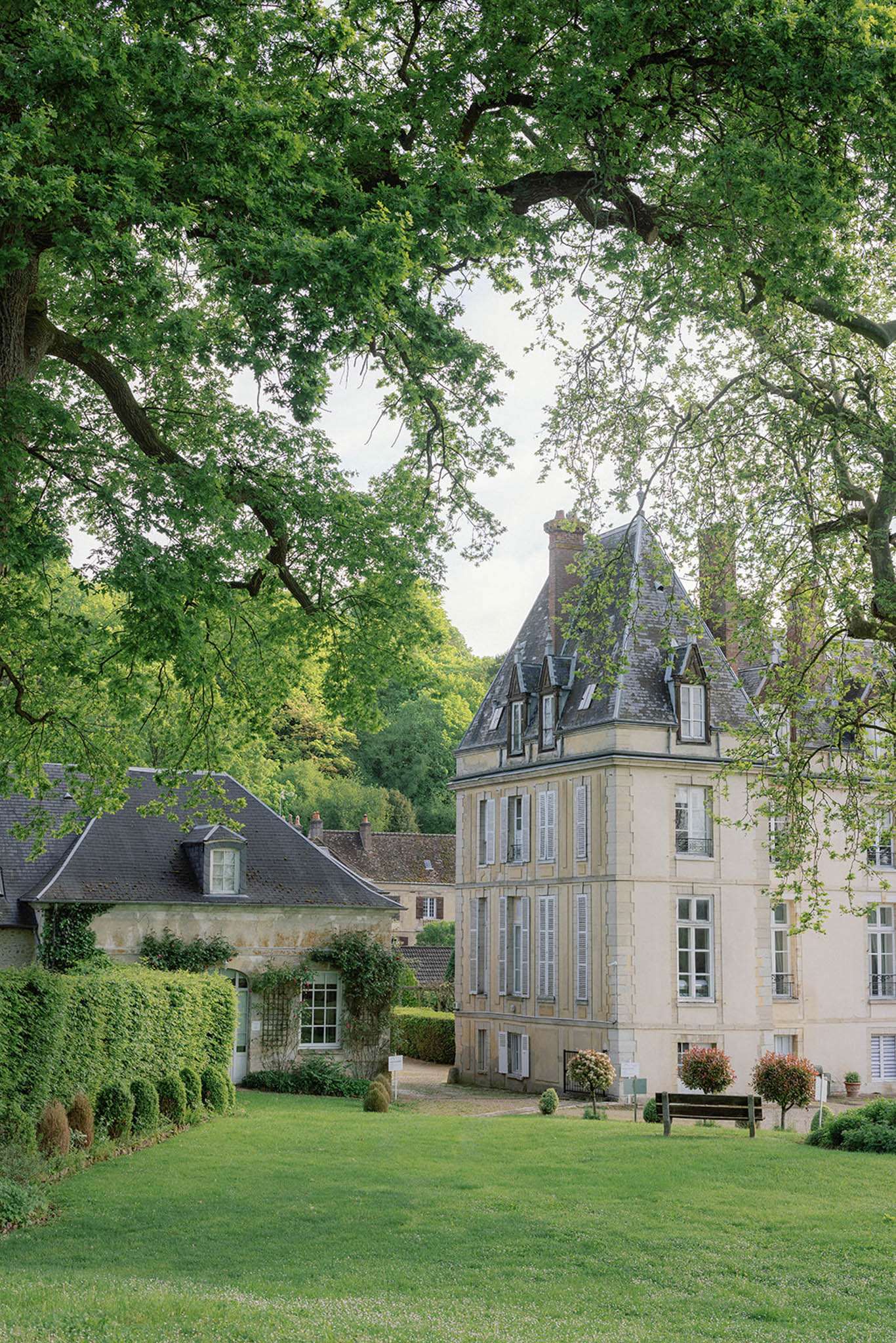 A wide exterior shot of a French château and its grounds, with no people visible. The main building features cream-colored stone facades, blue-grey slate mansard roofs with dormer windows, and white shuttered windows across three stories, including a prominent corner tower. To the left, a smaller stone outbuilding with climbing plants and a pitched slate roof sits behind a trimmed hedge border with rounded topiary accents. The foreground shows a well-maintained lawn, and a wooden bench is positioned near the château entrance alongside small ornamental trees with rounded canopies. The composition is shot from ground level across the open grounds, framed by large mature tree branches overhead. Potential venue feature image.