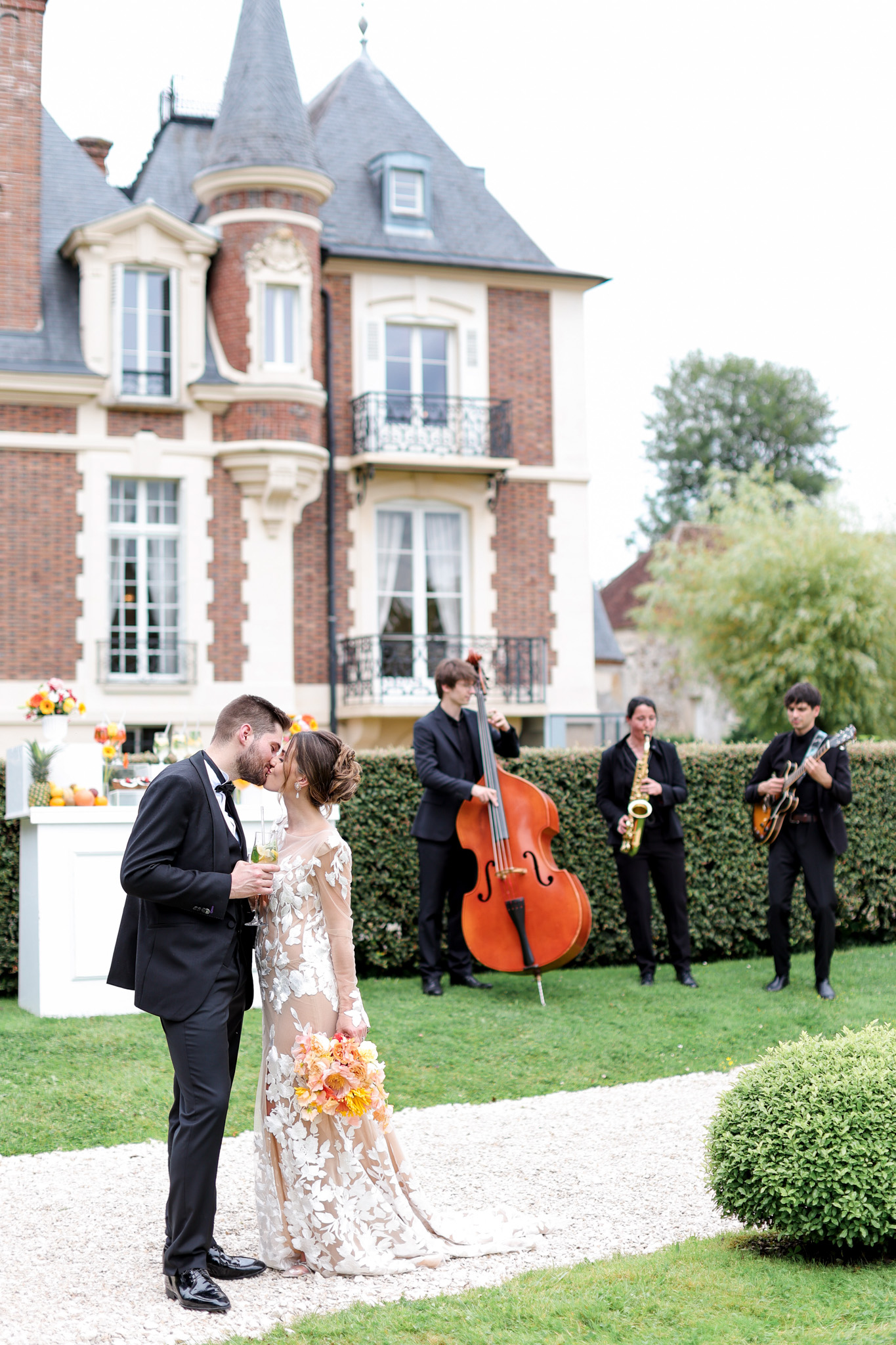 Couple kisses before red-brick manor as jazz trio plays double bass, saxophone, and guitar at cocktail hour