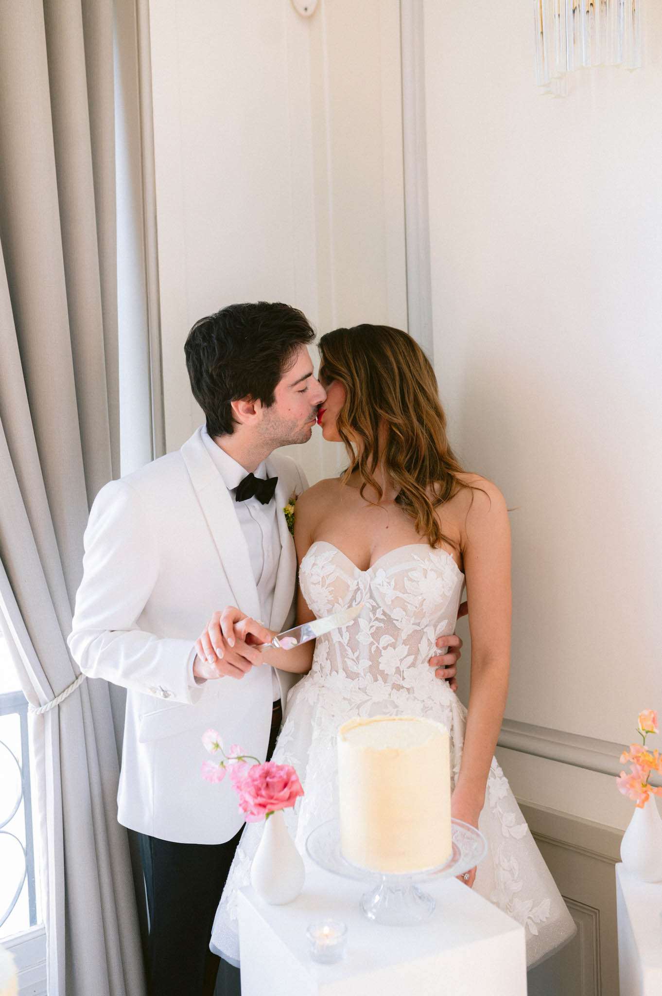 The bride and groom share a kiss during the cake cutting moment inside a bright, white-walled interior room with tall grey drape curtains and a crystal wall sconce visible in the background. The groom wears a white dinner jacket with black bow tie and black trousers, while the bride wears a strapless sweetheart-neckline lace gown with floral appliqué detailing and a nude underlayer. They hold a cake knife together in front of a single-tier ivory cake with a subtle gold-edged top, displayed on a white pedestal cake stand. Small white bud vases holding bright pink roses and peach-toned florals are arranged around the cake table, adding pops of color against the all-white setting. The overall styling is clean and modern with a white-and-pink color palette. Portrait-style mid shot.
