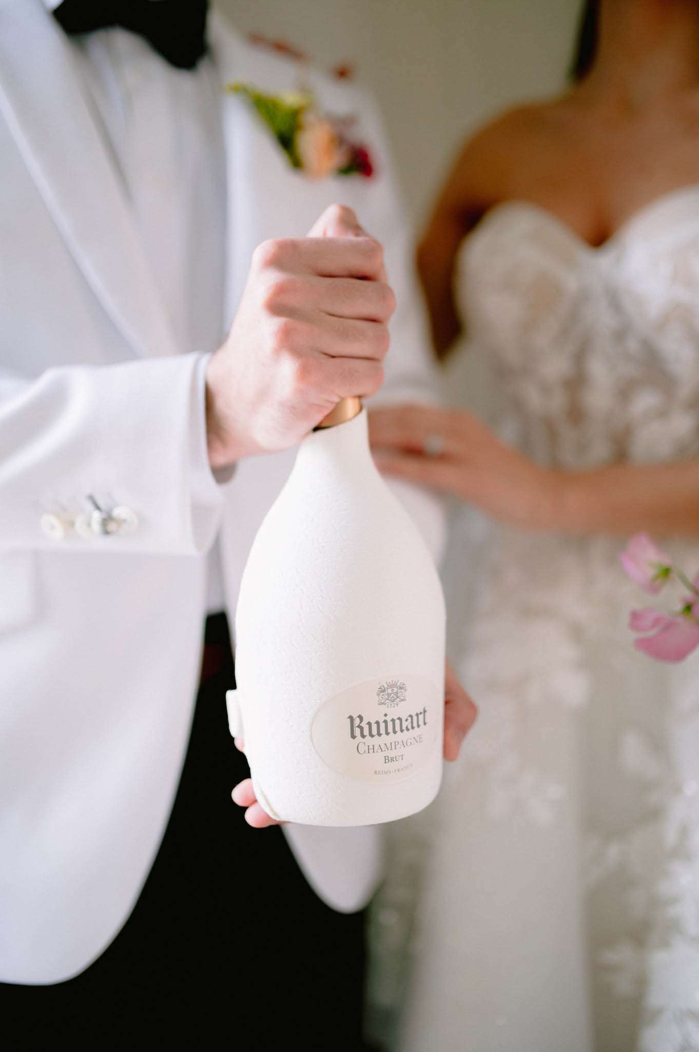 Close-up detail shot of a couple holding a bottle of Ruinart Brut Champagne in its distinctive white second-skin eco-designed casing with a gold foil neck. The groom is wearing a white dinner jacket with a black bow tie and small white floral buttonhole, while the bride, partially visible in the background, is wearing a strapless lace wedding gown with floral appliqué detailing. Pink blooms are visible at the edge of the frame, likely part of the bride's bouquet. The composition is tightly cropped on the champagne bottle as the focal point, with the couple softly blurred behind it.