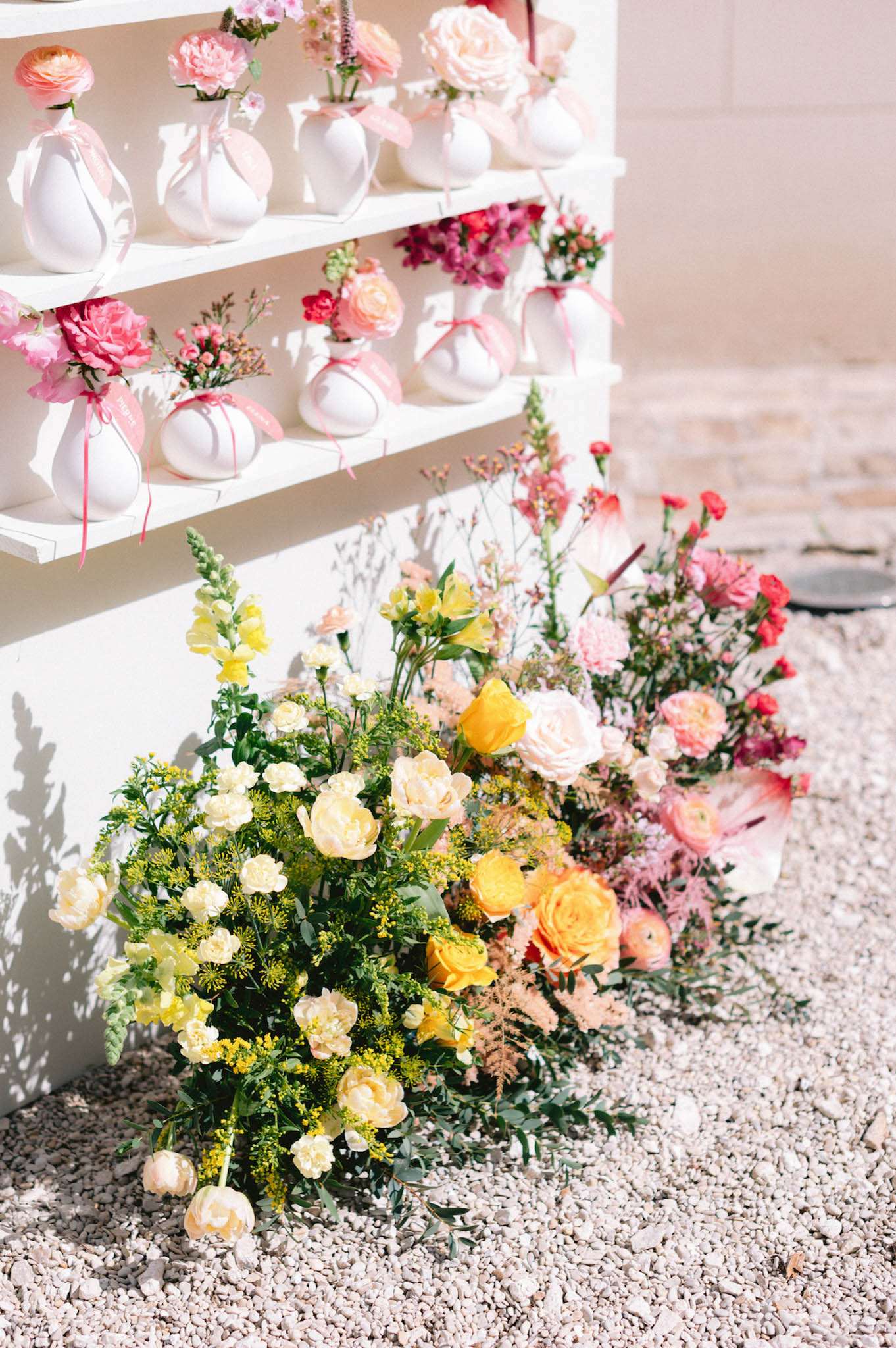 A floral decor display photographed outdoors on a gravel surface, showing a white painted wooden shelving unit holding approximately 20 small round white ceramic bud vases, each tied with a pink satin ribbon and containing individual blooms including ranunculus, carnations, garden roses, and small filler flowers in a palette of hot pink, blush, and coral. At the base of the shelving unit, two large ground-level floral arrangements are placed directly on the gravel: one on the left featuring cream and yellow peonies, white carnations, yellow snapdragons, and chartreuse filler foliage, and one on the right featuring coral and hot pink carnations, pink ranunculus, and blush astilbe, with the two arrangements transitioning through peach and orange garden roses in the center. The overall floral palette moves from yellow and cream on the left to deep pink and coral on the right, suggesting a gradient color scheme. The styling is garden-party inspired with a bright, summery aesthetic. This is a close-up detail shot taken in bright natural light.