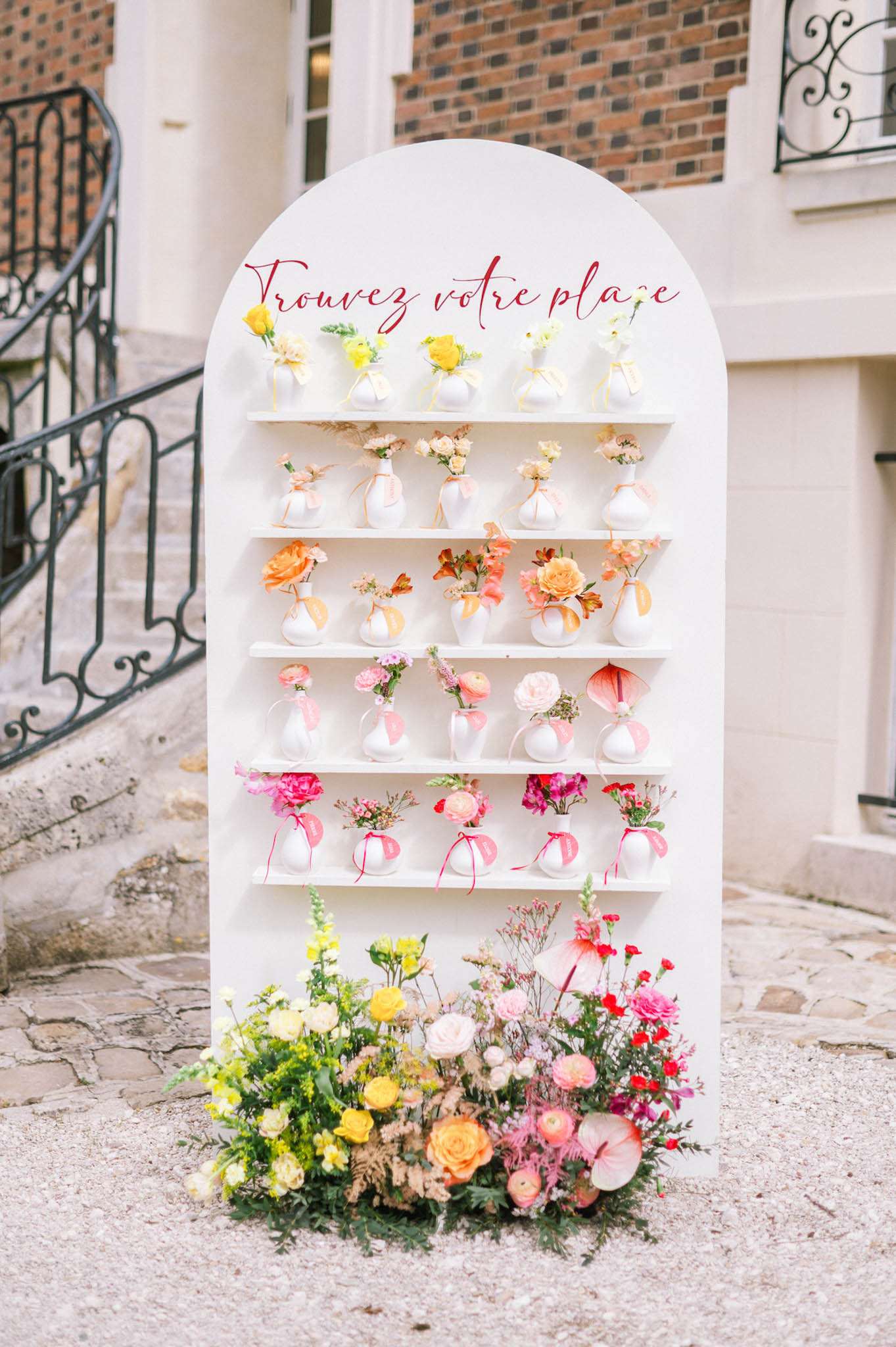 A wedding seating chart display photographed outdoors in front of a brick and stone building with a wrought-iron staircase railing. The display is a white arch-shaped panel with five horizontal shelves, each holding small white ceramic bud vases containing individual flower arrangements in a gradient color palette — yellow roses and ranunculus at the top, transitioning through peach and coral garden roses, soft pink ranunculus, hot pink carnations and flowers, down to the bottom shelves. Each vase is tied with a coordinating satin ribbon in matching colors. The header reads 'Trouvez votre place' in deep red calligraphy script. The base of the display is flanked by two loose floral ground arrangements featuring yellow, orange, peach, and hot pink blooms including roses, ranunculus, carnations, and anthurium. The overall floral palette is vibrant and multicolored, with a modern yet playful styling approach. Wide, slightly elevated portrait-orientation shot showing the full display in context.