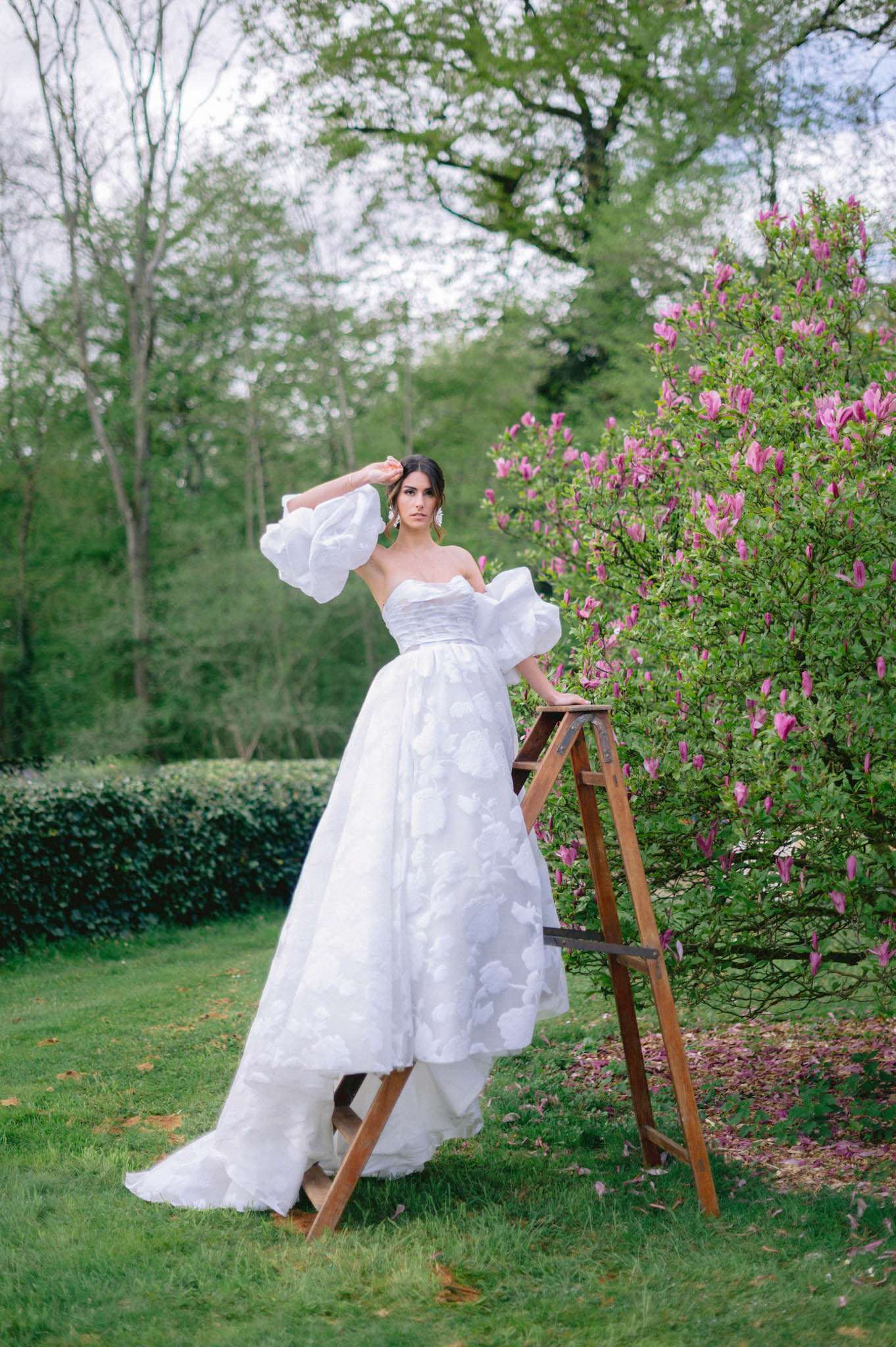 Bride on rustic ladder beside pink magnolia in white ballgown with dramatic organza puff sleeves