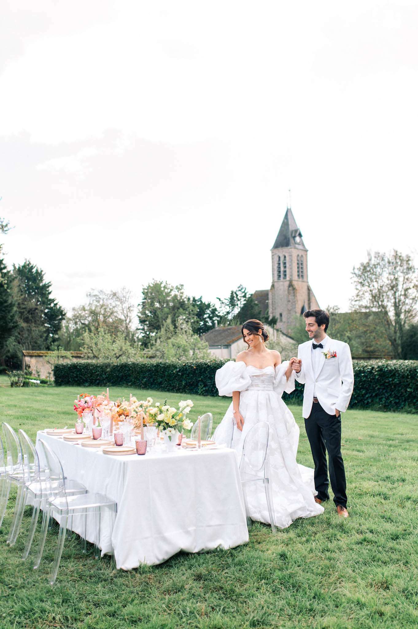 Bride in puff-sleeve ballgown and groom in white jacket beside table with coral and orange floral runner