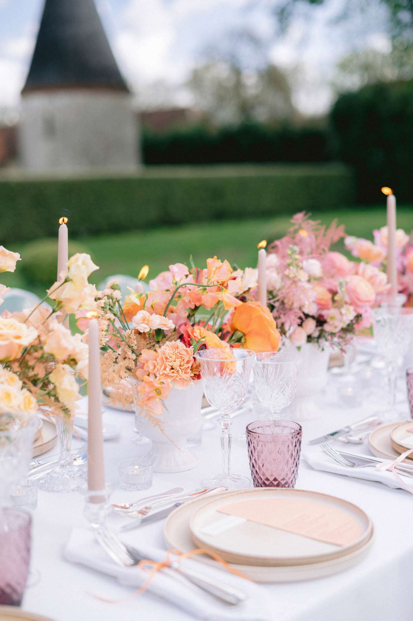 Peach ranunculus and blush sweet pea centerpiece in milk-glass compotes with mauve tumblers and lit tapers