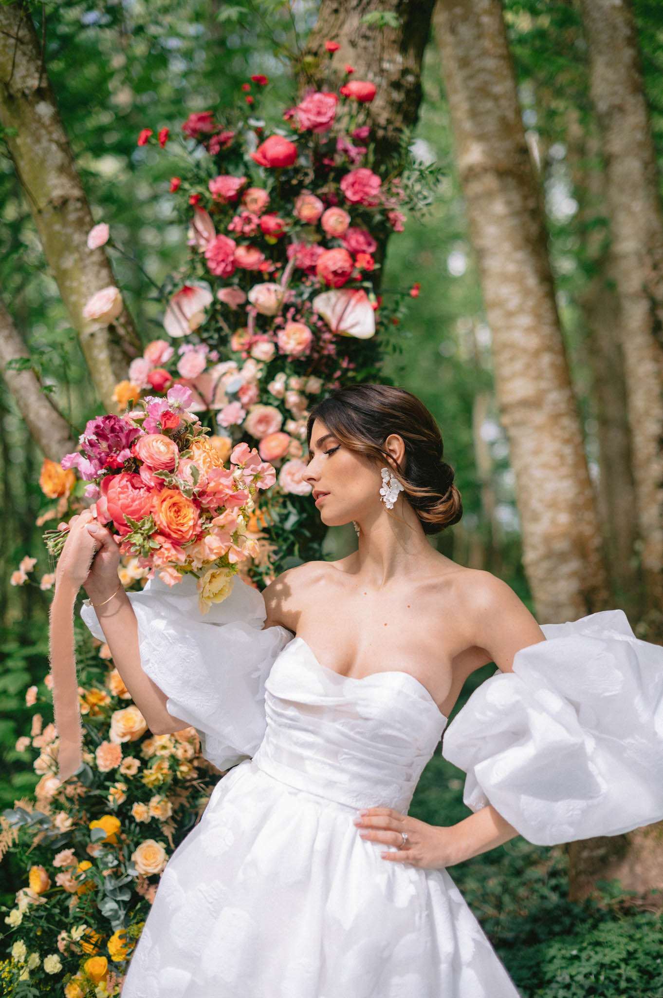 Bride in puff-sleeve ballgown with coral peony bouquet beside gradient pink-to-yellow floral tree wrap