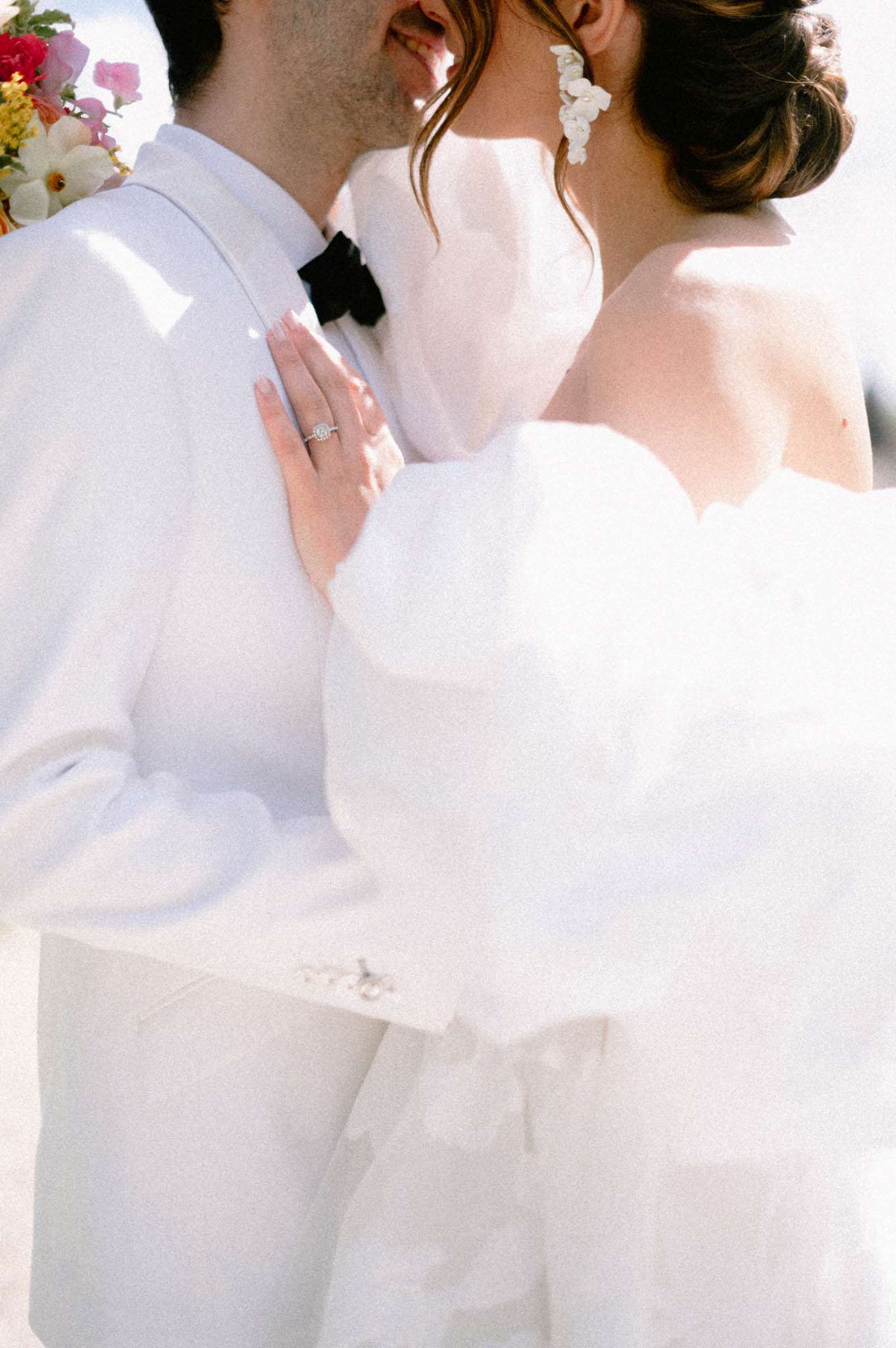 Close-up couple chest level bride's hand showing halo diamond ring on groom's white jacket with floral bouquet