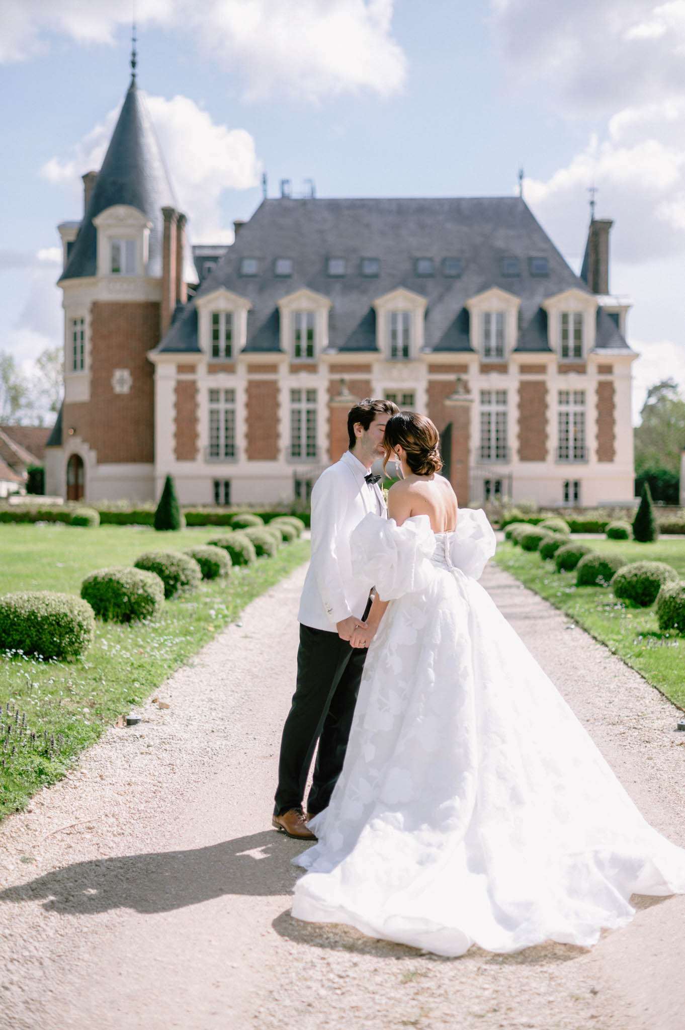 Bride and groom kissing on gravel path leading to French chateau with mansard roof and turret