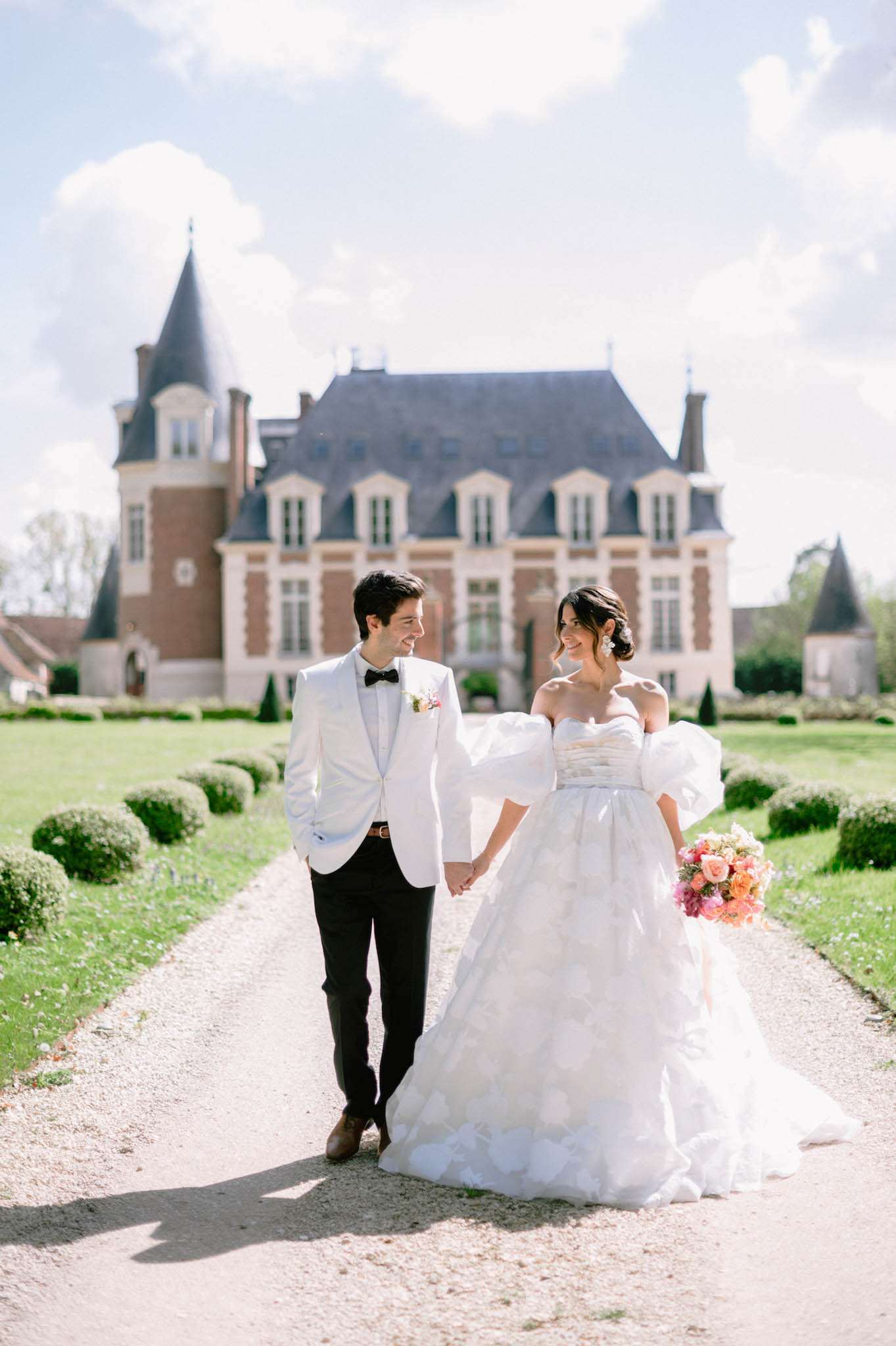 A couple portrait taken outdoors on a gravel path leading toward a French château, with a formal parterre garden featuring trimmed box hedges visible on either side. The bride wears a white strapless ballgown with a textured petal-layered skirt and detachable oversized puff sleeves, paired with statement drop earrings and an updo; she carries a lush bouquet of coral, peach, and pink roses. The groom wears a white dinner jacket with black bow tie, black trousers, a tan leather belt, and a small floral boutonnière in pink and white tones. The couple walks hand in hand, looking at each other and smiling, in a mid-length portrait composition with the château's red brick and stone facade, mansard roof, and conical tower visible softly in the background.