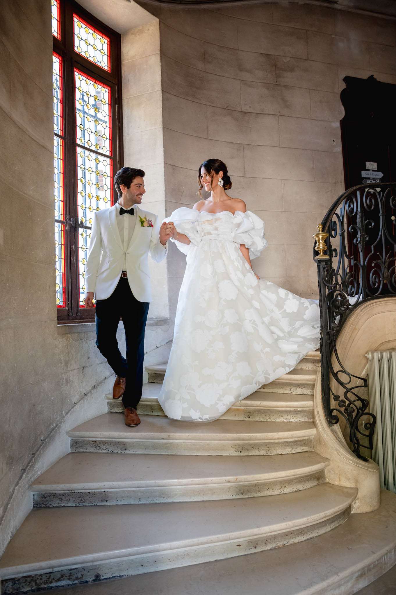 A couple portrait taken indoors on a grand curved stone staircase, likely inside a French château or historic mansion. The couple is holding hands and descending the stairs while looking at each other and smiling. The bride wears an off-the-shoulder white ball gown with large puff sleeves and an all-over floral appliqué pattern across the full skirt, paired with statement drop earrings and an updo hairstyle. The groom wears a white dinner jacket with a black bow tie, navy trousers, and brown leather dress shoes, with a small pink and white boutonnière on his lapel. The staircase features a wrought-iron railing with ornate scrollwork and a brass newel post, and a tall stained-glass window with red, yellow, and clear panes floods the landing with natural light. The overall styling is classic and formal. Wide portrait shot.