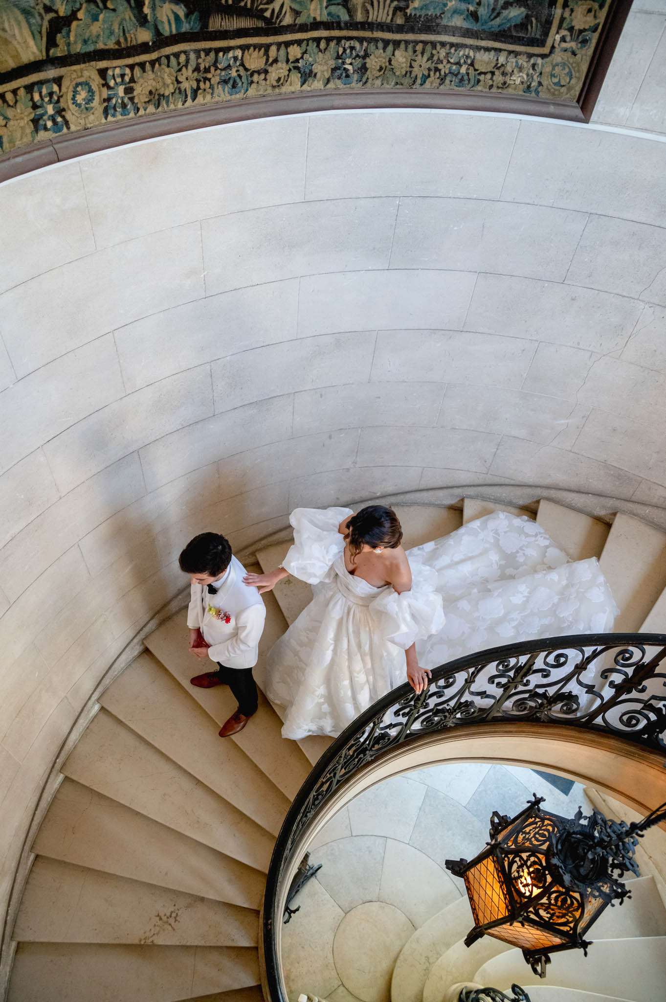 Overhead view of bride in puffed-sleeve ballgown and groom descending a limestone spiral staircase in a chateau