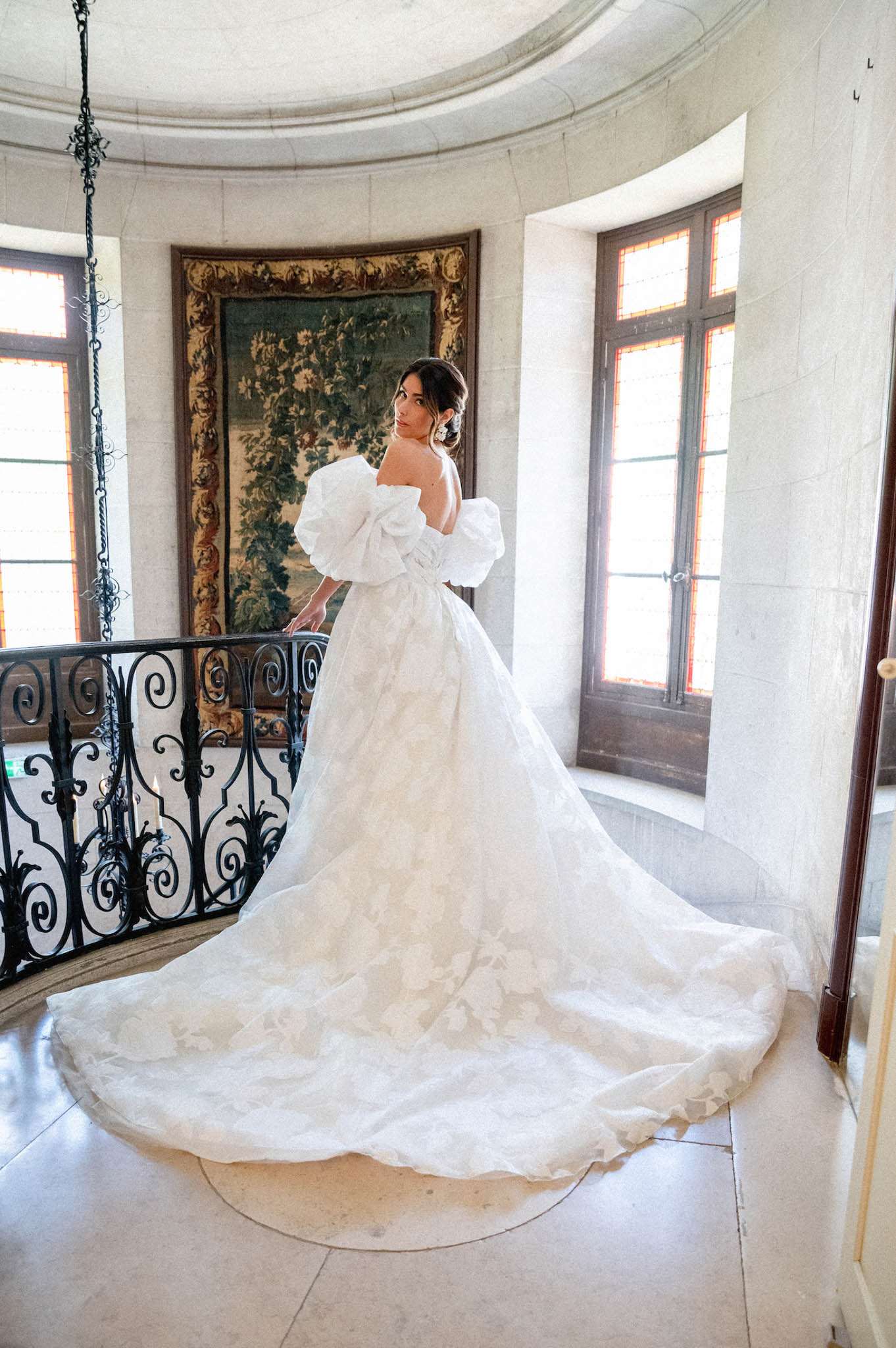 Bride in off-the-shoulder puff-sleeve ball gown with long train looks over her shoulder on a chateau staircase landing