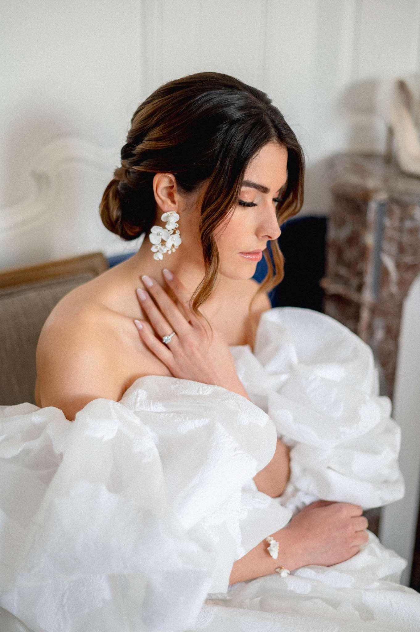 A close-up portrait of a bride seated indoors, likely in a French château room with white panelled walls and an ornate marble fireplace visible in the background. She is wearing an off-the-shoulder white gown with voluminous ruched organza sleeves and detachable puff sleeves, styled in a classic modern aesthetic. Her dark hair is worn in a low chignon with a loose face-framing tendril, and she wears statement white floral drop earrings with pearl accents, a matching floral bracelet cuff, and a solitaire engagement ring with a blue stone. The shot is a medium portrait with soft, bright natural light emphasizing the bridal accessories and gown texture.