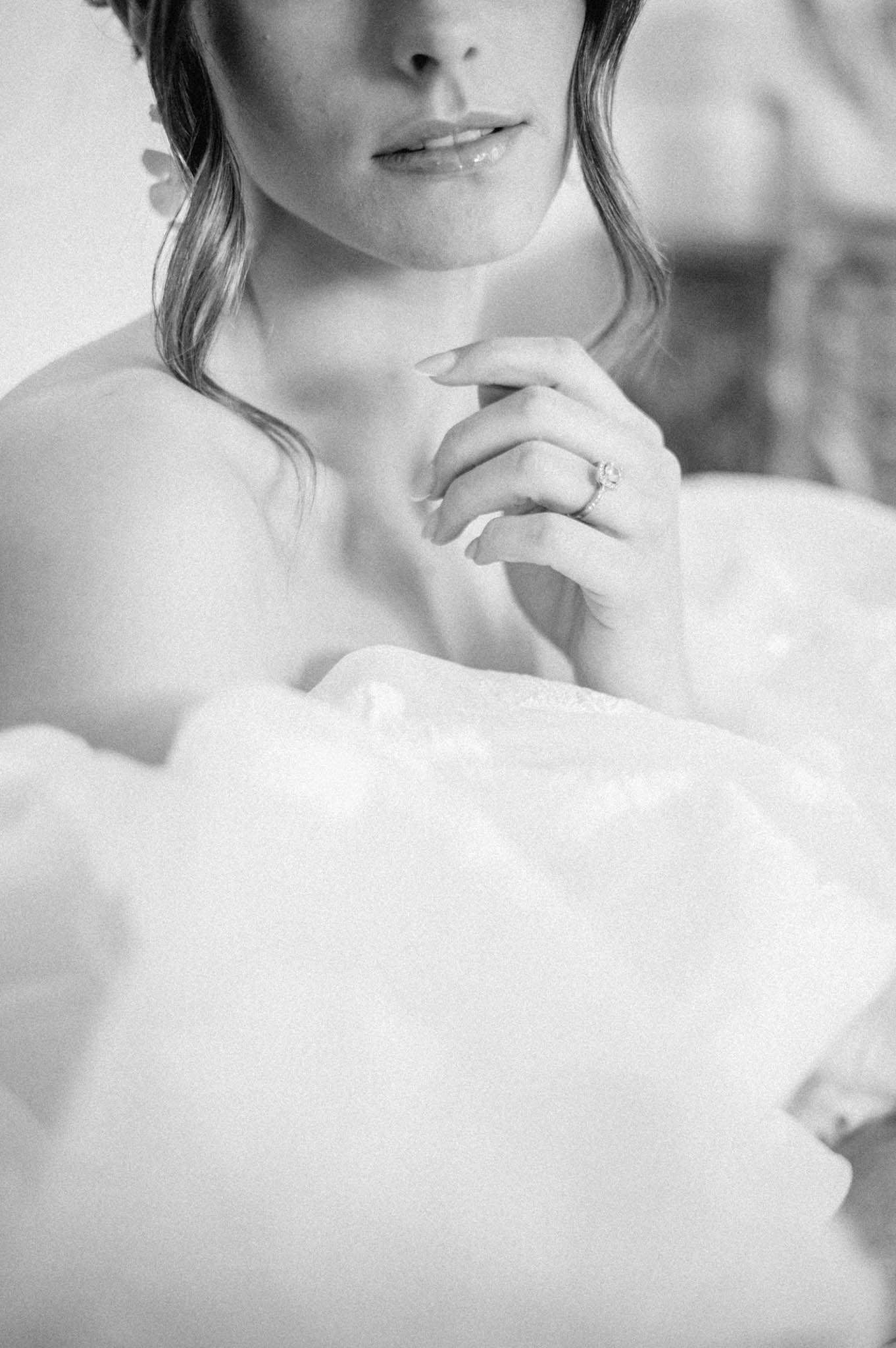 A black-and-white close-up portrait of a bride sitting in a bubble bath, captured from the chin down to mid-torso. The frame is filled with soft, voluminous foam bubbles in bright whites, while the bride's bare shoulders and décolletage are visible above them. Her hand is raised to her chest, prominently displaying a diamond solitaire engagement ring with a pavé band. Her hair is loosely styled and she appears to wear a small floral hair accessory. The image has a soft, fine-grain film quality with gentle contrast and bright highlights in the foam. This is a getting-ready or bridal boudoir-style detail shot.