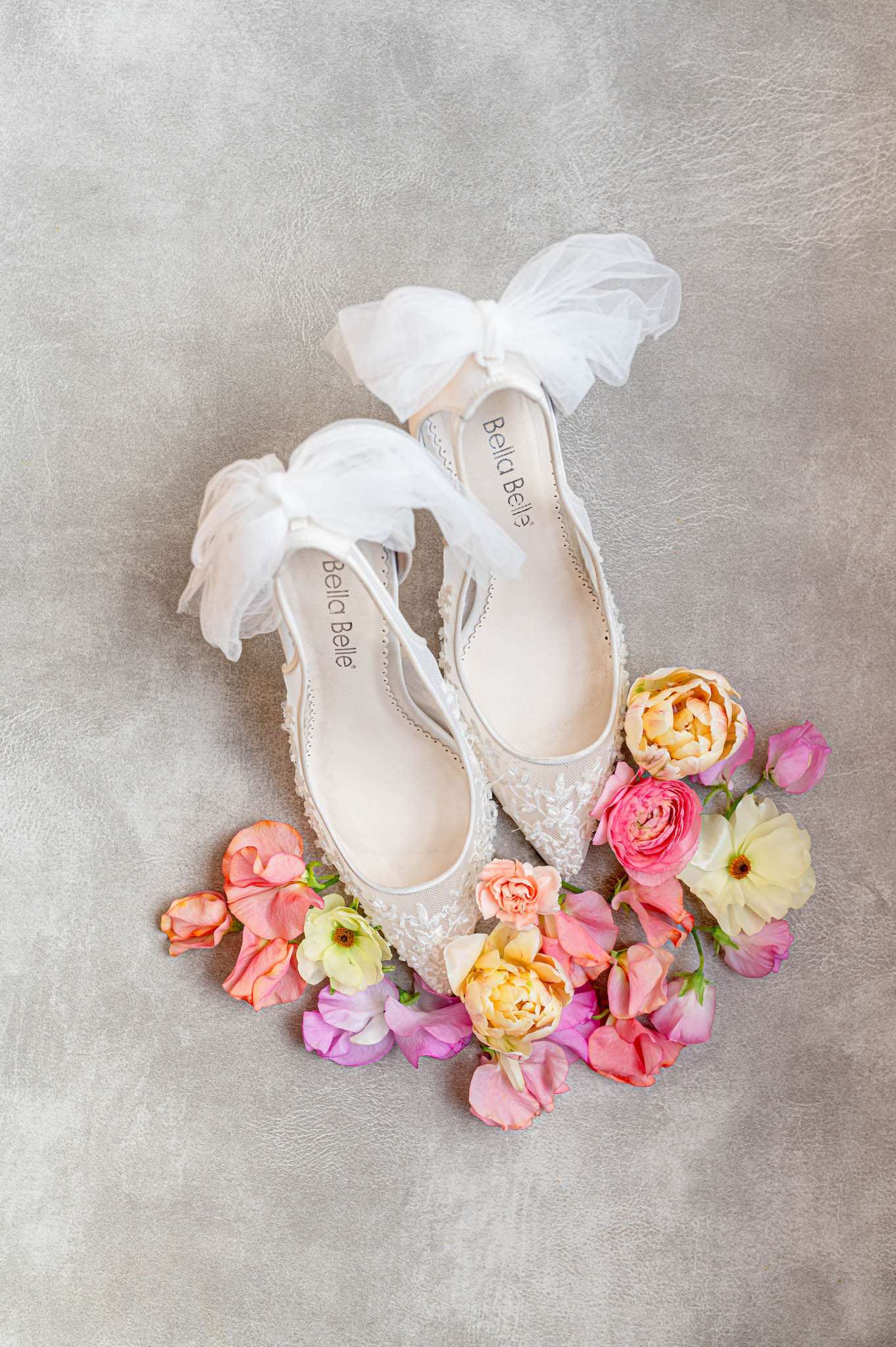 Bella Belle ivory lace bridal flats with tulle bows surrounded by peach ranunculus, sweet peas, and coral tulip petals