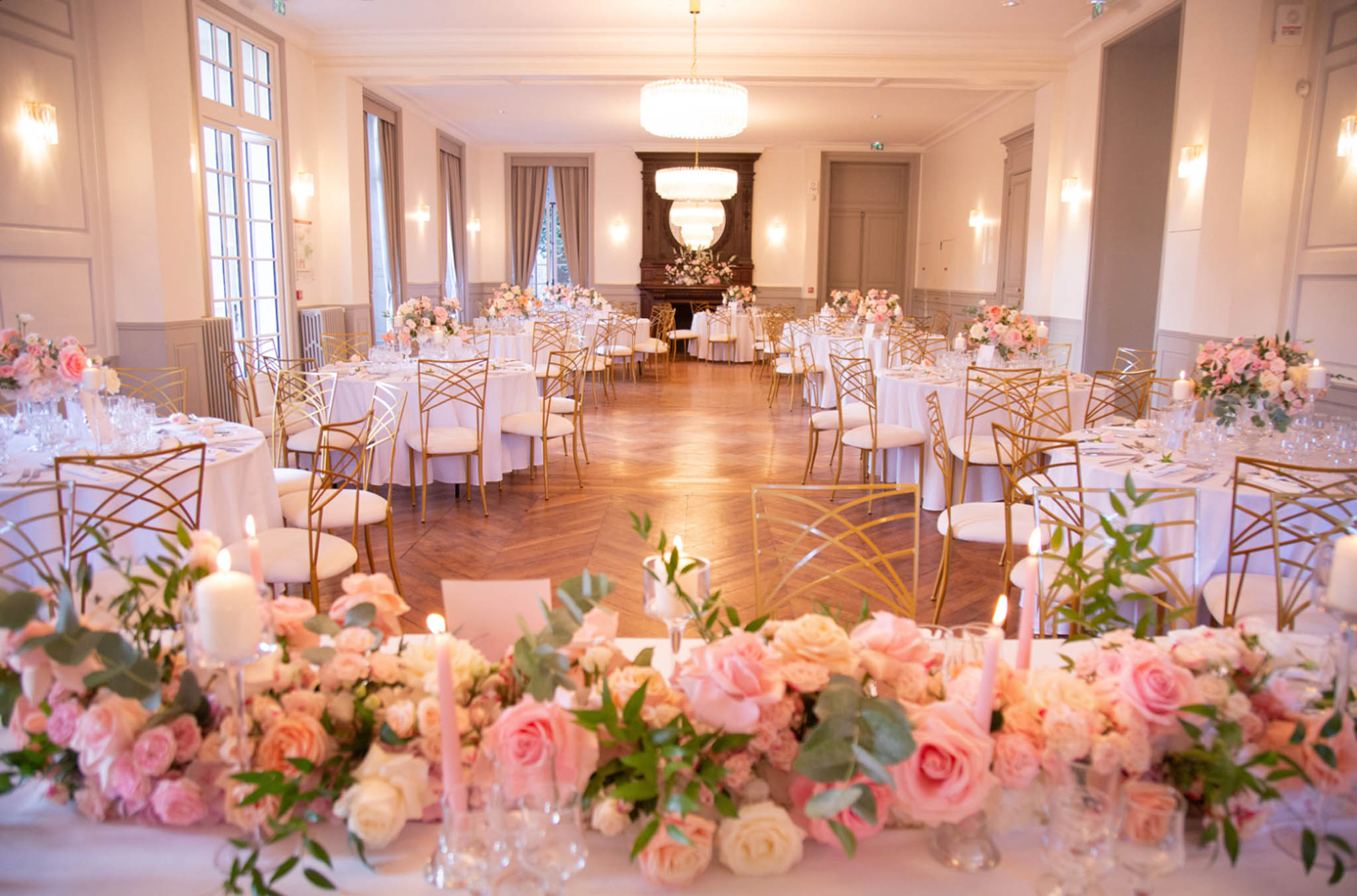 A wide shot of an indoor wedding reception ballroom set up for a seated dinner, with no guests present. The room features herringbone parquet flooring, white-painted panelled walls with wall sconces, grey wainscoting, tall French windows with grey draping curtains, a dark wood fireplace at the far end, and two tiered crystal chandeliers overhead. Round guest tables are dressed in white floor-length linens and surrounded by gold geometric-frame chairs with white cushioned seats. Centrepieces on the guest tables consist of blush pink and cream floral arrangements. In the foreground, a long head table is decorated with a dense runner of blush pink, coral, and ivory roses with eucalyptus greenery, interspersed with lit white pillar candles and slim blush pink taper candles in clear glass holders. The overall decor palette is blush pink, ivory, and gold, in a classic French interior style. Potential venue feature image.