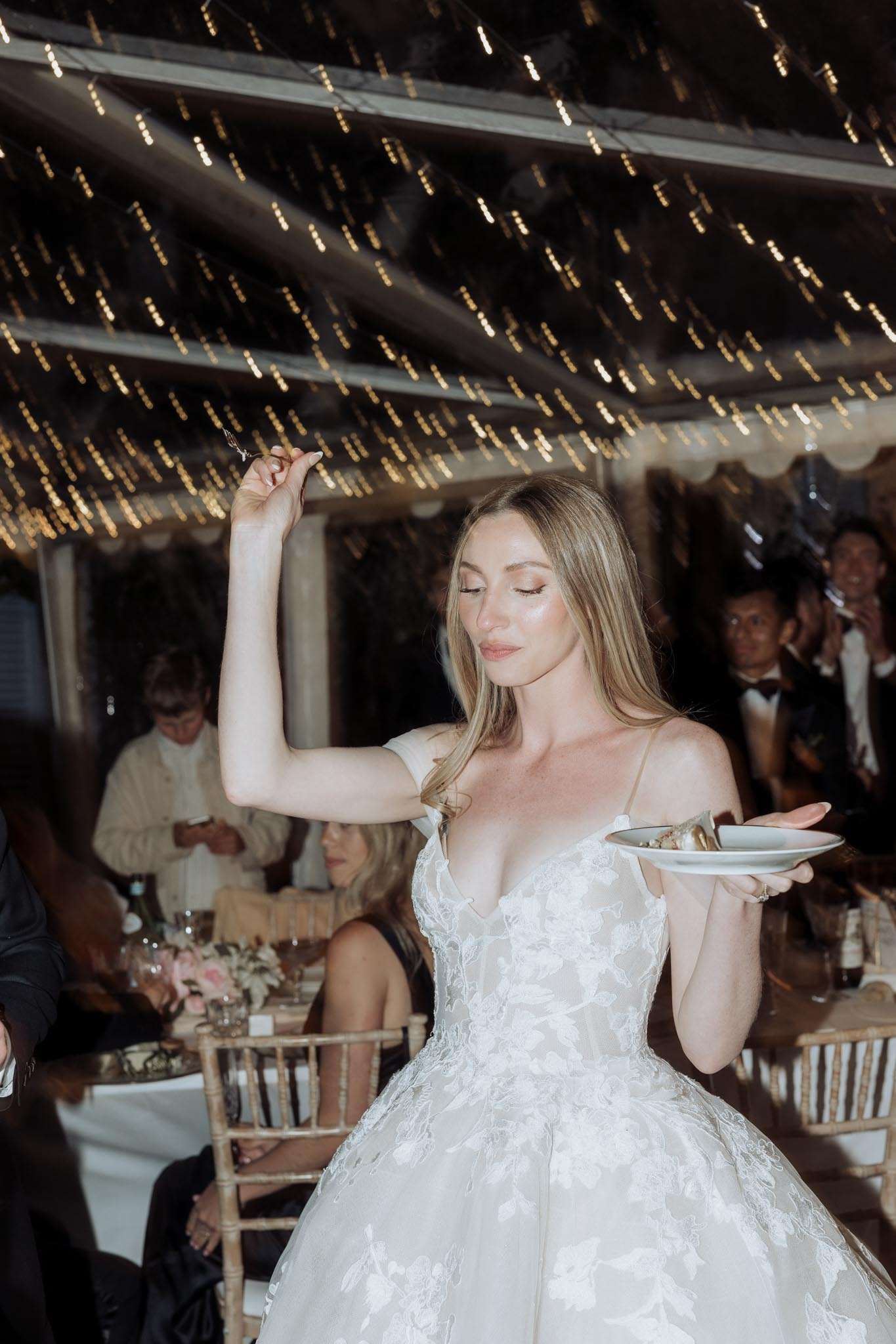 Bride eating cake in floral lace ballgown under fairy-light canopy with gold Chiavari chairs behind