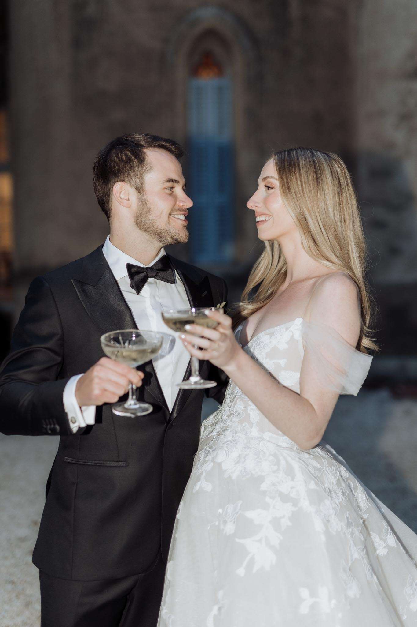 A couple shares a champagne toast outdoors in front of what appears to be a historic stone building with Gothic-arched windows visible in the soft-focus background. The groom wears a black tuxedo with satin lapels, white dress shirt, and black bow tie with silver cufflinks, while the bride wears an off-the-shoulder ivory ball gown with floral lace appliqué and a sheer tulle overlay on the sleeves. Both are holding coupe-style champagne glasses and are smiling at each other. This is a mid-shot couples portrait with a classic black-and-white formal styling aesthetic.