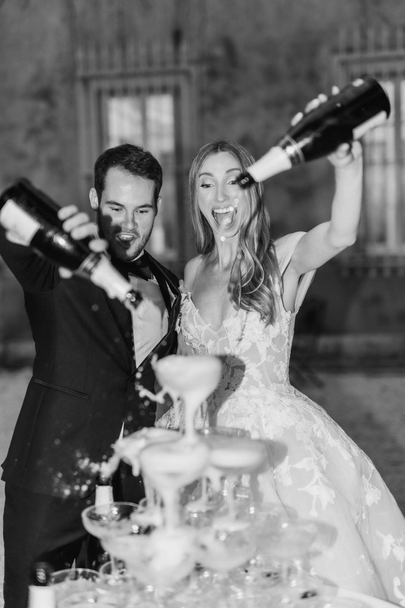 Black-and-white portrait-style shot of a bride and groom pouring champagne onto a coupe glass tower during their wedding reception. The groom wears a black tuxedo with a bow tie and pours a bottle from his left side, while the bride raises a second bottle above her head with visible excitement, her mouth open and laughing. The bride wears a floral lace ballgown with thin straps and a deep V-neckline, with her hair worn down in loose waves. The champagne tower in the foreground features stacked coupe glasses with liquid overflowing down the tiers. The background is softly out of focus, showing what appears to be an outdoor setting against a stone building facade with an iron gate. The image has high contrast with bright highlights on the couple's faces and dress against a dark background.