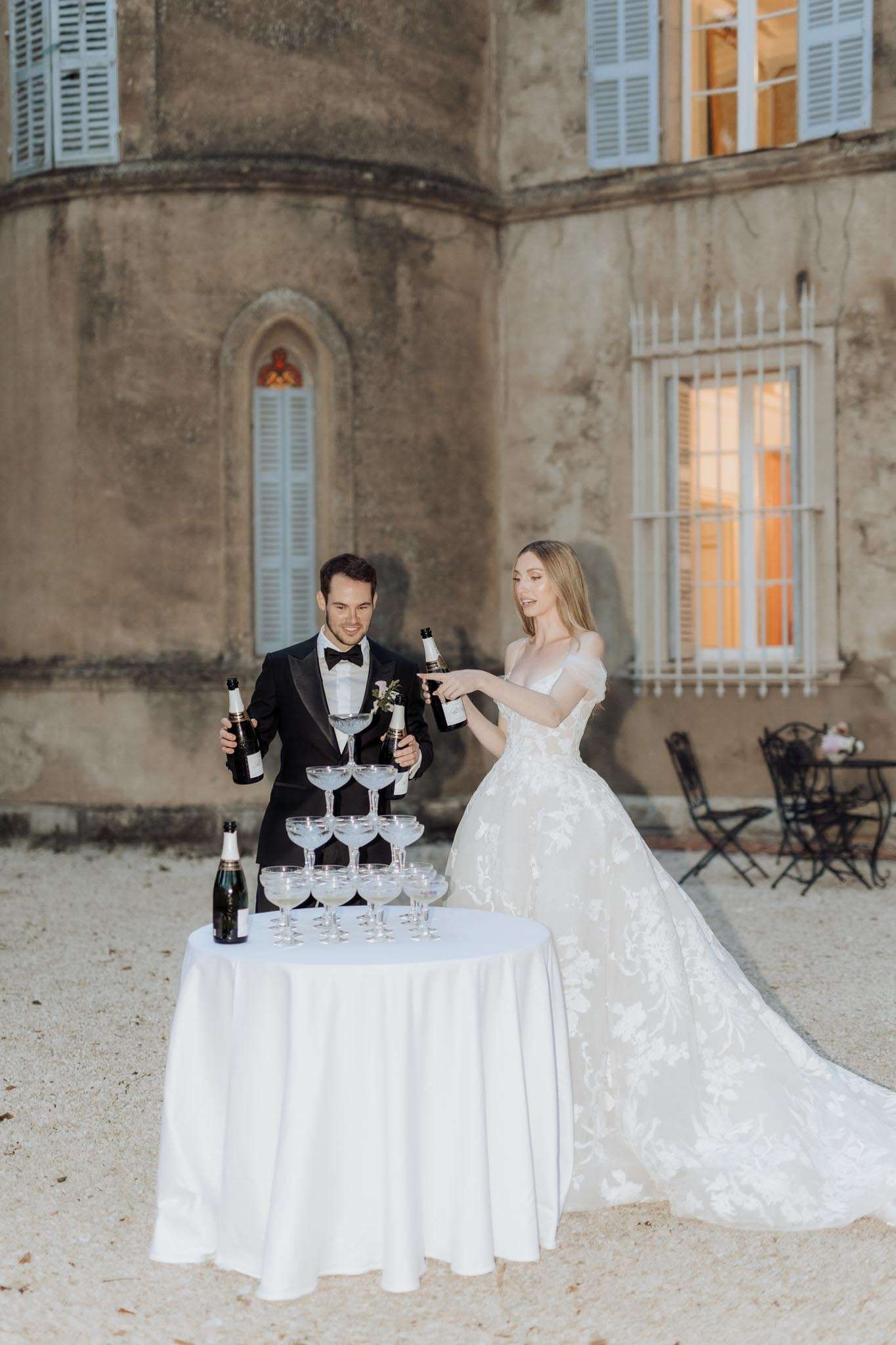 Bride and groom pouring champagne into coupe glass tower in chateau courtyard at dusk