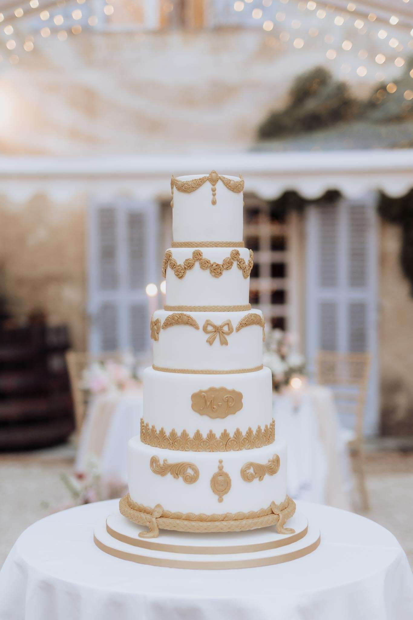 Five-tier white fondant wedding cake with gold baroque sugar-work decorations and monogram plaque