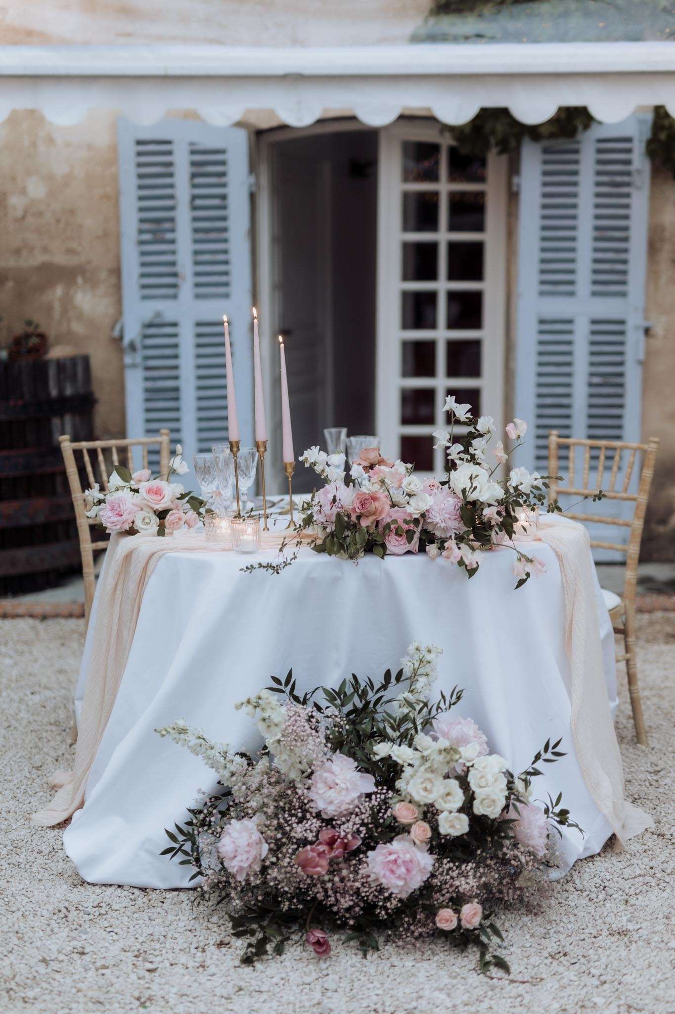 Sweetheart table with blush runner, gold chiavari chairs, taper candles, and rose garland on gravel courtyard