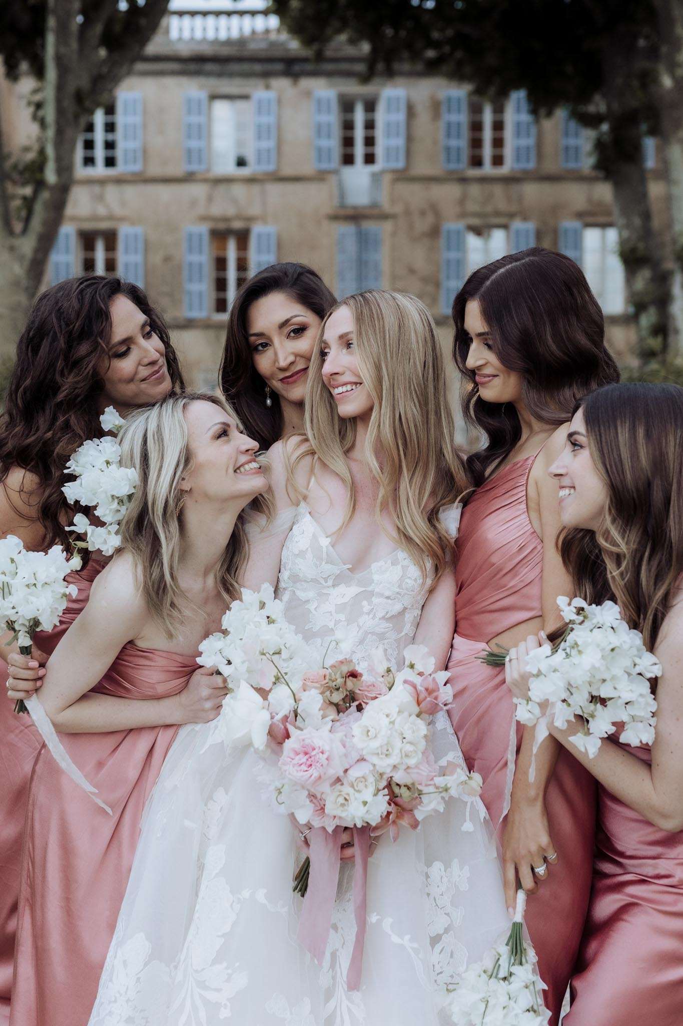 Bride with five bridesmaids in dusty rose and mauve satin dresses holding sweet pea bouquets before chateau