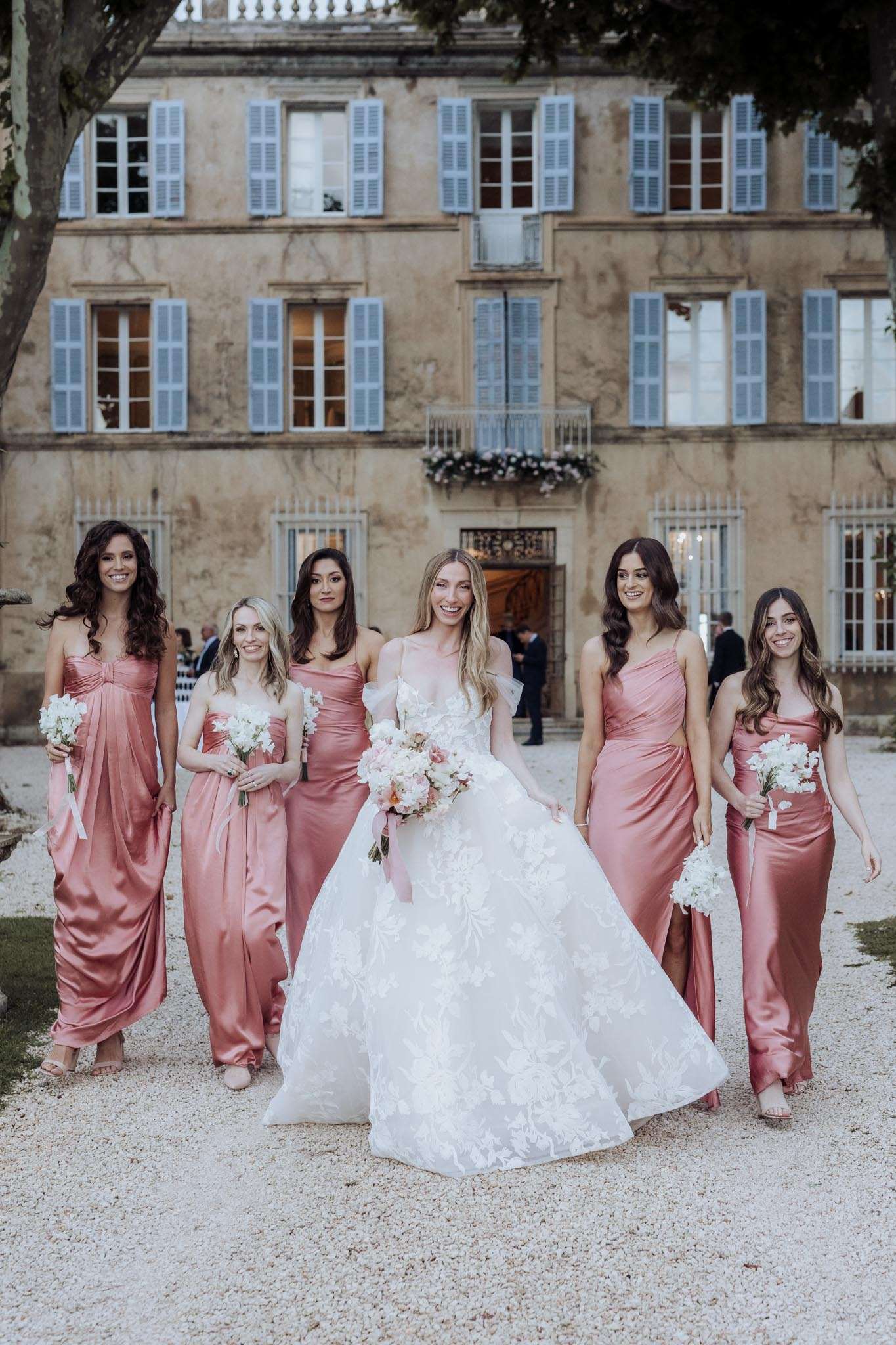Bride in off-shoulder ball gown with five bridesmaids in dusty rose satin dresses walking on chateau courtyard
