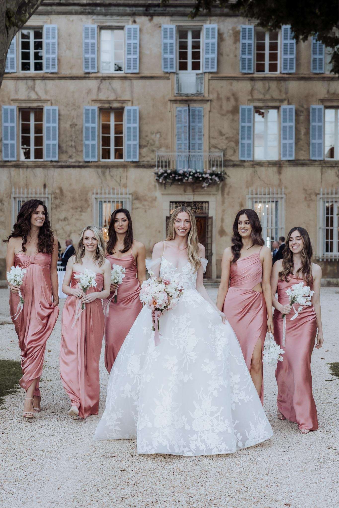 A bridal party portrait taken outdoors on a gravel forecourt in front of a large French château with pale ochre stone walls and powder blue shutters. The bride stands at center, wearing a full ball gown with an off-the-shoulder tulle ruffle neckline and a floral-appliqué skirt, carrying a bouquet of blush peonies, white blooms, and a trailing dusty pink ribbon. She is flanked by five bridesmaids wearing mismatched satin rose-pink floor-length gowns in varying silhouettes — draped, strapless, and one-shoulder styles — each holding a bouquet of white sweet peas or similar white blooms. A floral arrangement of pink and white flowers is visible decorating a balcony railing on the château facade behind the group. The shot is a mid-distance walking portrait with the full group in frame, conveying a classic French château wedding aesthetic with a warm rose-pink color palette.