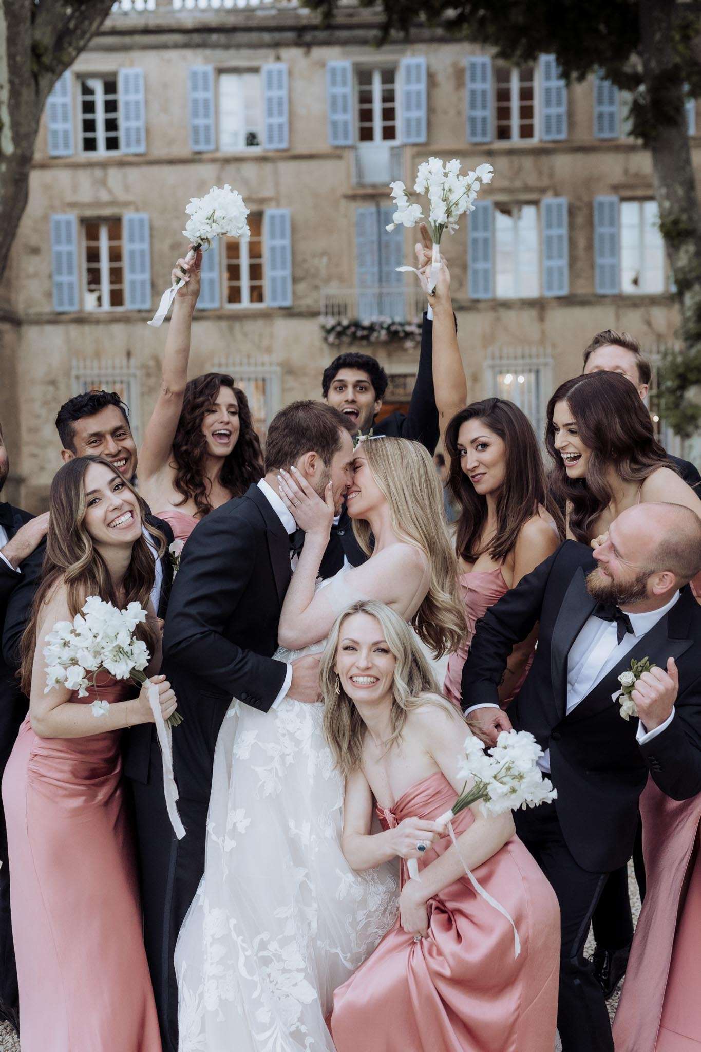 Couple kisses amid cheering bridesmaids in dusty rose and groomsmen in black tie before blue-shutter chateau