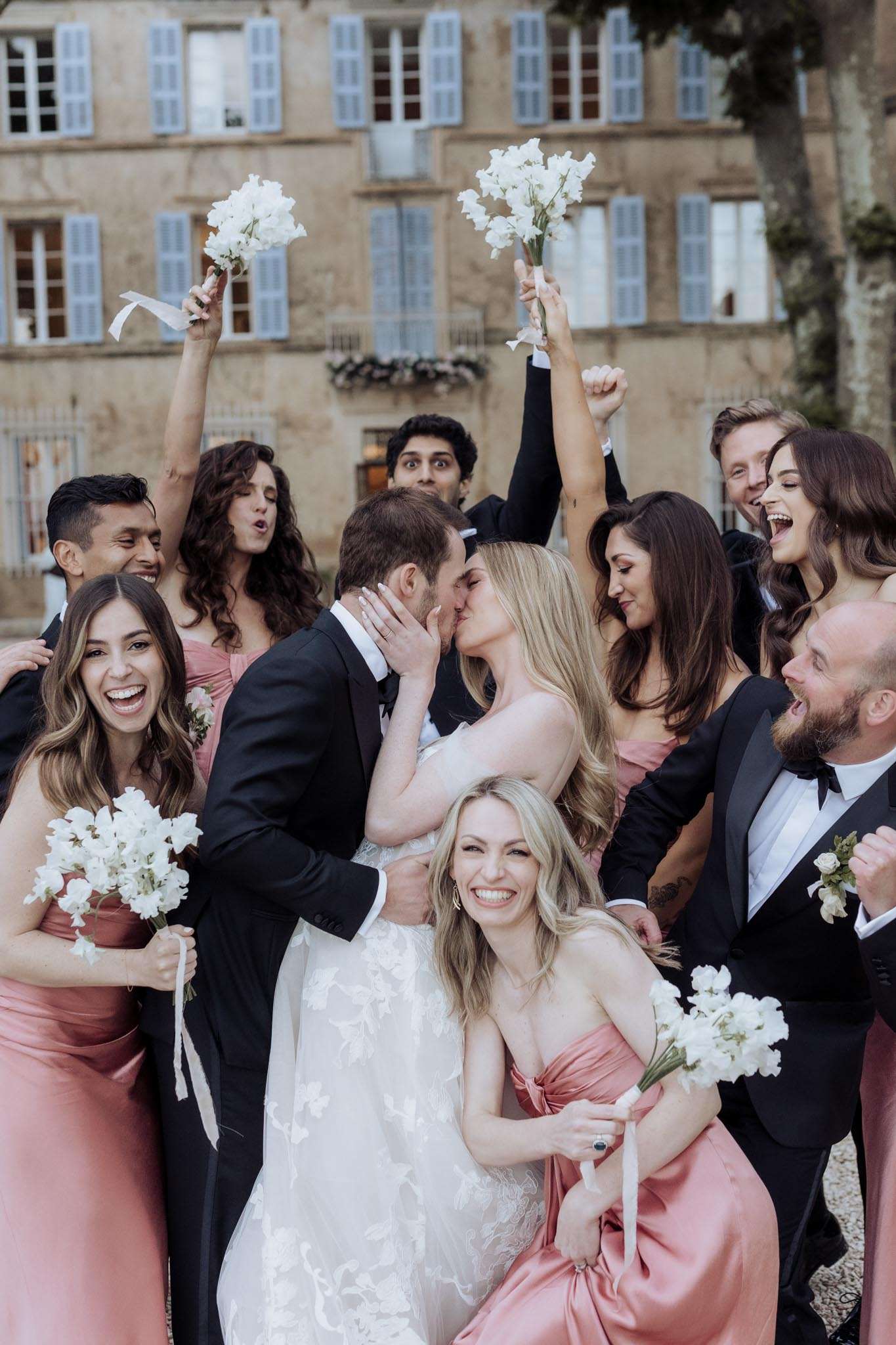 Bridal party of ten cheering as couple kisses, bridesmaids in dusty rose gowns before blue-shuttered chateau