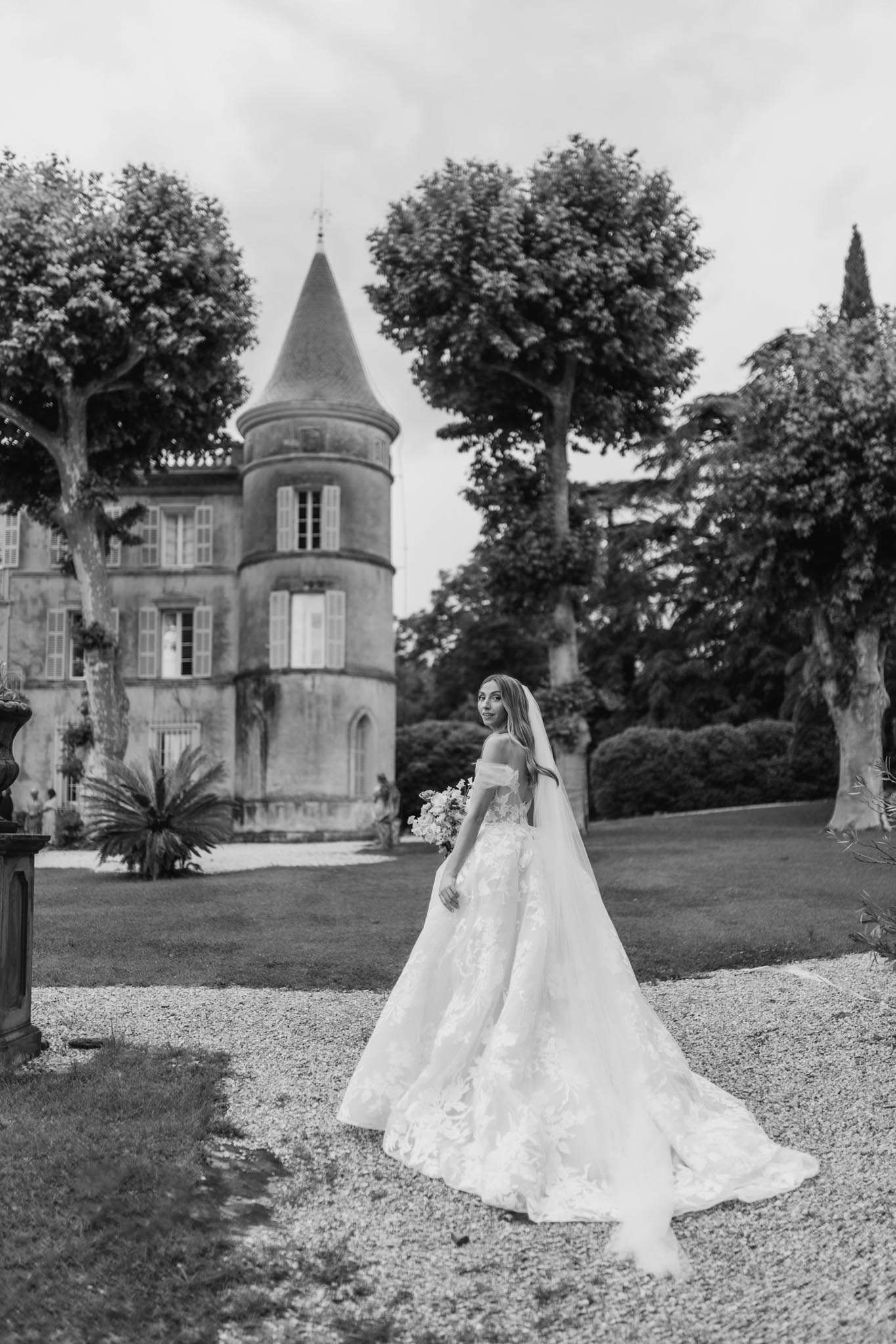 This is a black-and-white bridal portrait taken outdoors on the grounds of a French château featuring a distinctive round tower with a conical roof and shuttered windows. The bride stands on a gravel path and looks back over her shoulder toward the camera, wearing an off-the-shoulder ball gown with heavy floral lace appliqué and an extended cathedral train spread across the gravel. She holds a mid-sized bouquet and wears a long veil falling behind her. The composition is a full-length portrait with the château centered in the background, framed by mature trees and manicured hedges. The image has high contrast with bright highlights on the dress against the darker mid-tones of the grounds and building. Potential venue feature image.