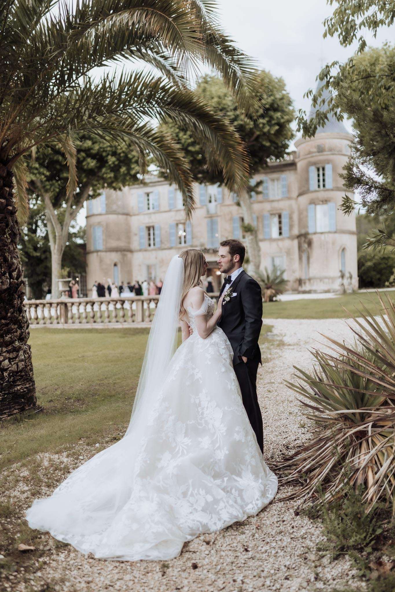 Bride in off-shoulder lace ball gown and groom in navy tuxedo near-kiss on chateau grounds with blue shutters