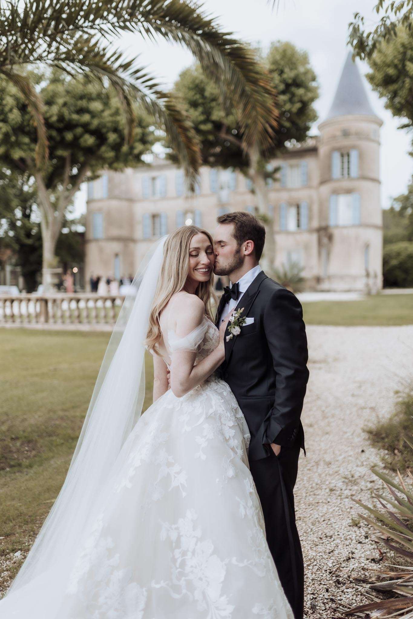 Groom kisses bride in floral applique ballgown with cathedral veil on gravel path with chateau turret behind