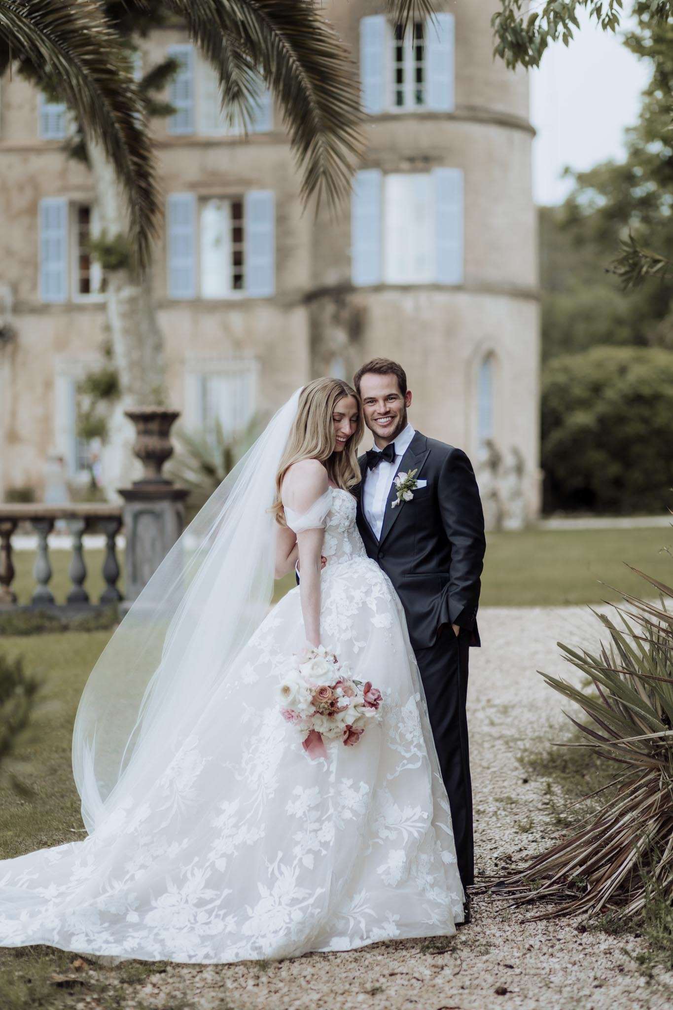 Bride in floral lace ballgown with veil and groom in navy tuxedo before blue-shuttered chateau tower