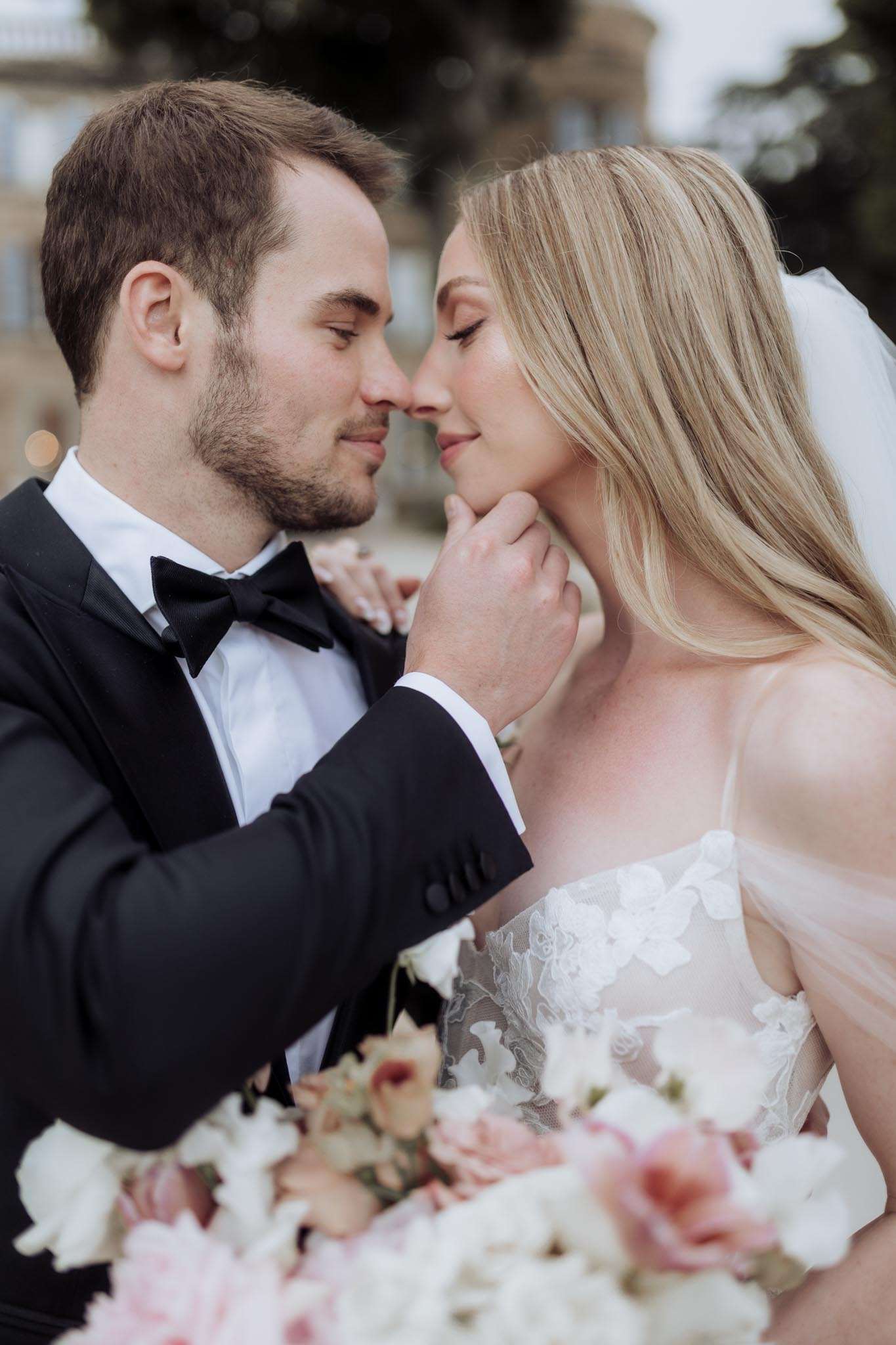 A close-up portrait of a bride and groom facing each other with their foreheads nearly touching, shot outdoors with a blurred château-style building visible in the background. The groom wears a black tuxedo with a white dress shirt and black bow tie, and gently cups the bride's chin with one hand. The bride wears a spaghetti-strap white lace and tulle gown with floral appliqué detailing and a long white veil, with her blonde hair worn loose. In the foreground, slightly out of focus, is a bridal bouquet featuring white blooms and blush pink roses. The overall styling is classic and formal, with a soft, warm color palette.