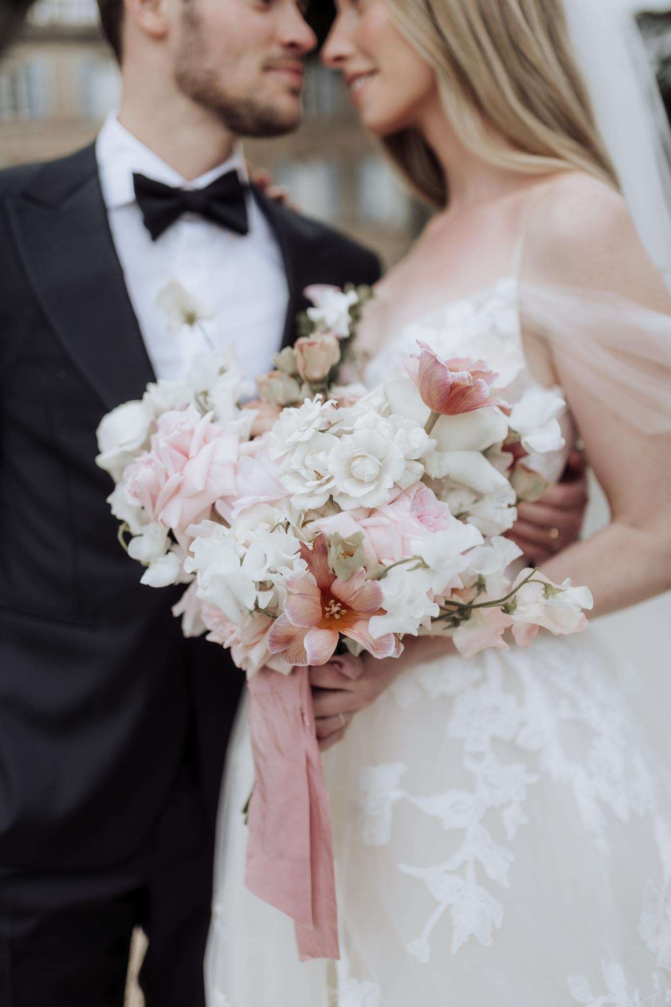 Close-up of bridal bouquet with blush tulips, coral amaryllis, and white ranunculus held by couple touching foreheads