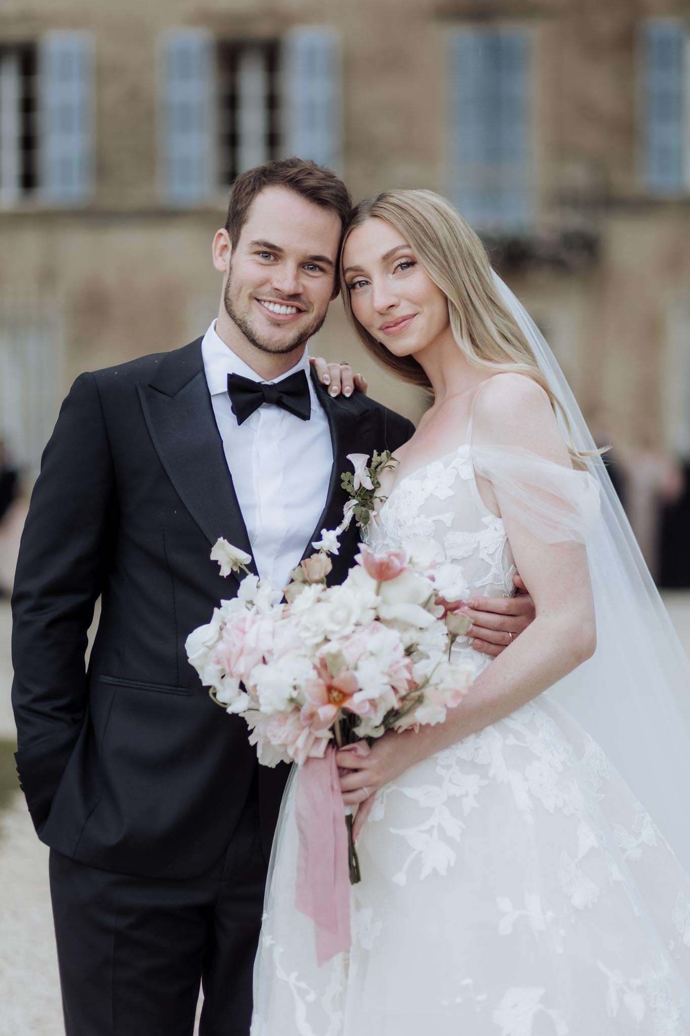 Bride in ivory lace ball gown and groom in black tuxedo posing in front of a French chateau