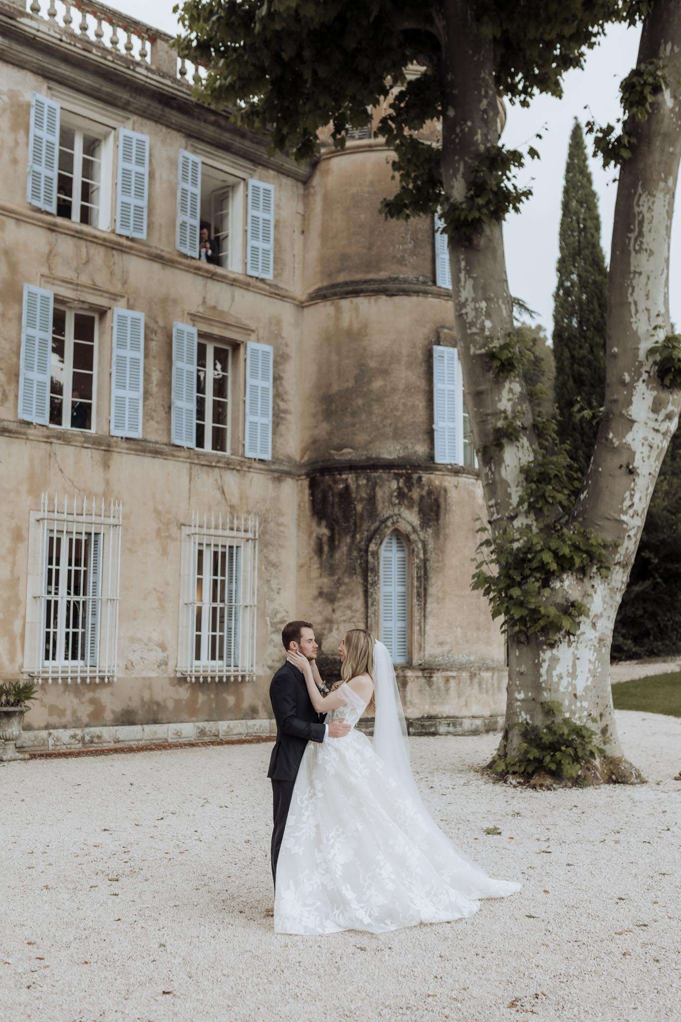 Bride in white lace ball gown with cathedral veil embracing groom in dark suit on gravel courtyard of French chateau