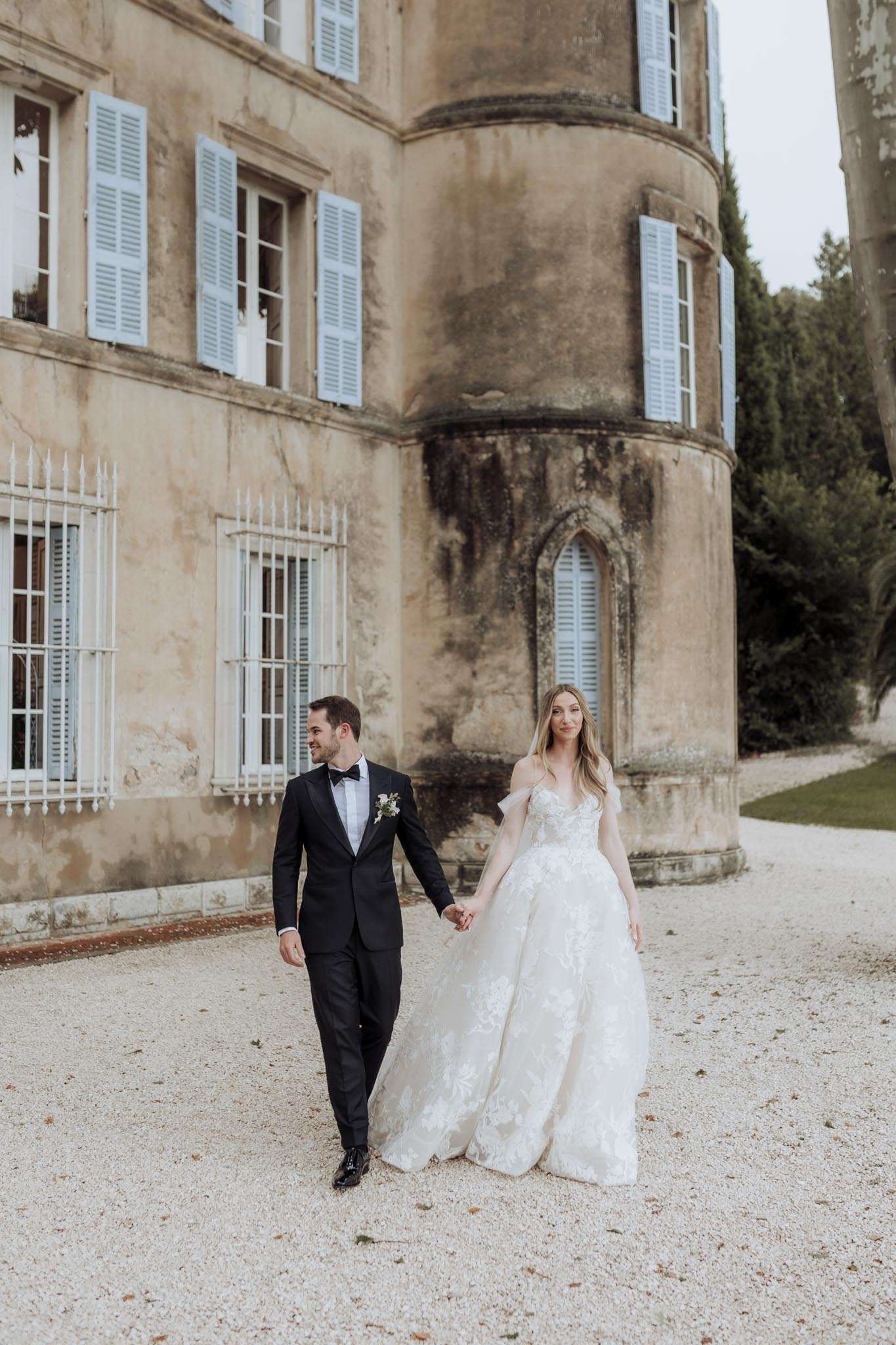 Couple walking on gravel before cylindrical stone tower with blue shutters bride in floral lace ballgown groom in tuxedo