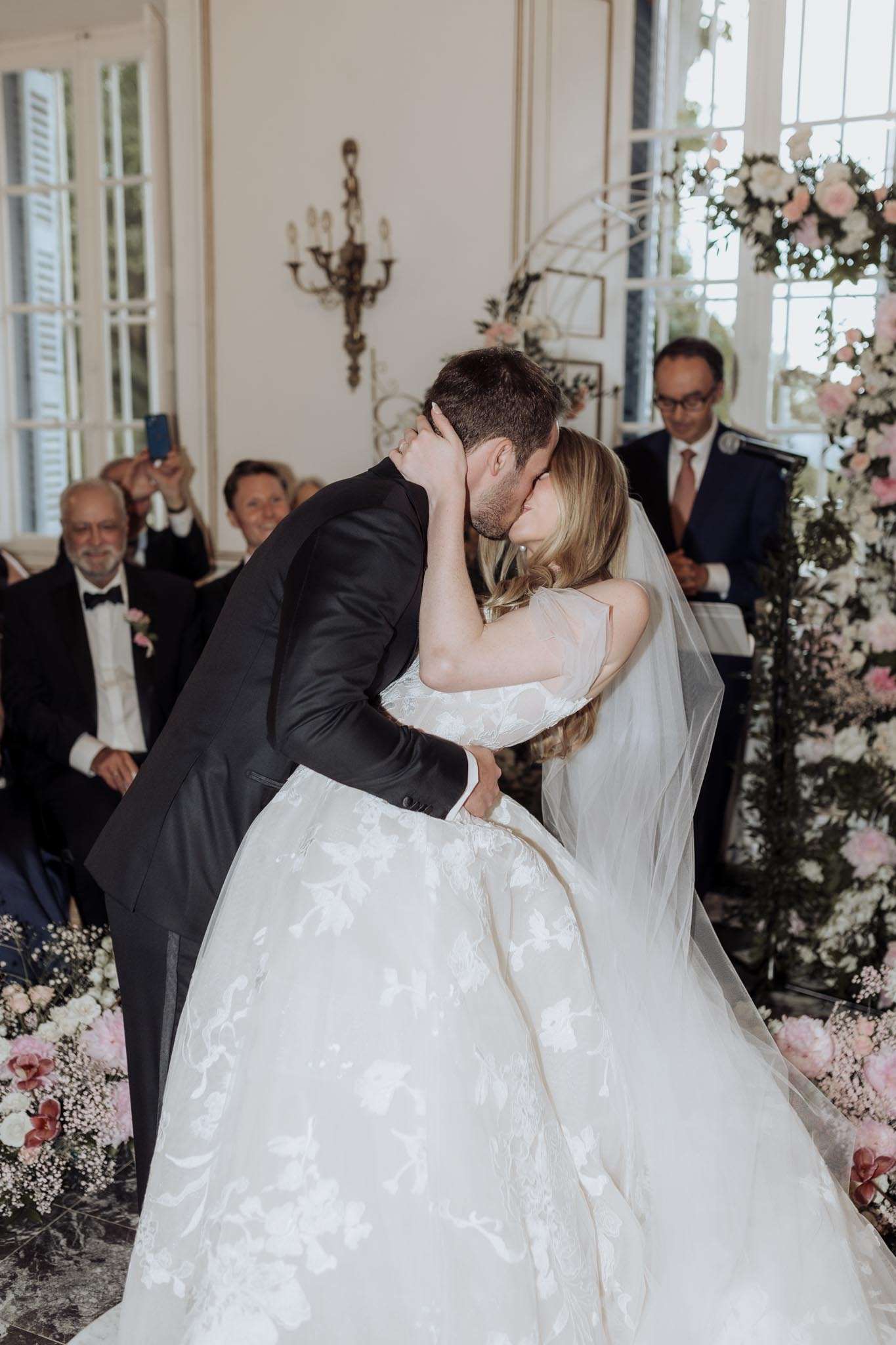 Bride and groom share first kiss under a blush and white floral arch in a chateau salon with gold sconces