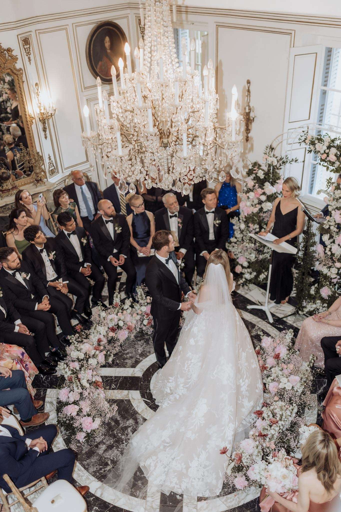 An indoor wedding ceremony captured from an elevated angle, showing the couple standing at the altar on a black and white marble floor, holding hands and facing each other. The bride wears a full-ballgown lace dress with a long cathedral veil and an extended train, while the groom is in a black tuxedo with a bow tie. A female officiant in a black dress stands at a lectern to the right. The ceremony space is decorated with abundant arrangements of blush pink peonies, white roses, gypsophila, and greenery lining the aisle floor on both sides, with a large floral arch of the same palette behind the officiant. Approximately 30 guests are seated on gold chiavari chairs on either side, many dressed in black tie attire. A large crystal and gold candelabra chandelier with lit white taper candles hangs prominently overhead, and the white paneled walls feature gold-framed mirrors and a portrait painting, consistent with a French château salon. The wide overhead shot captures the full scale of the ceremony setup and the bride's train spread across the marble floor.