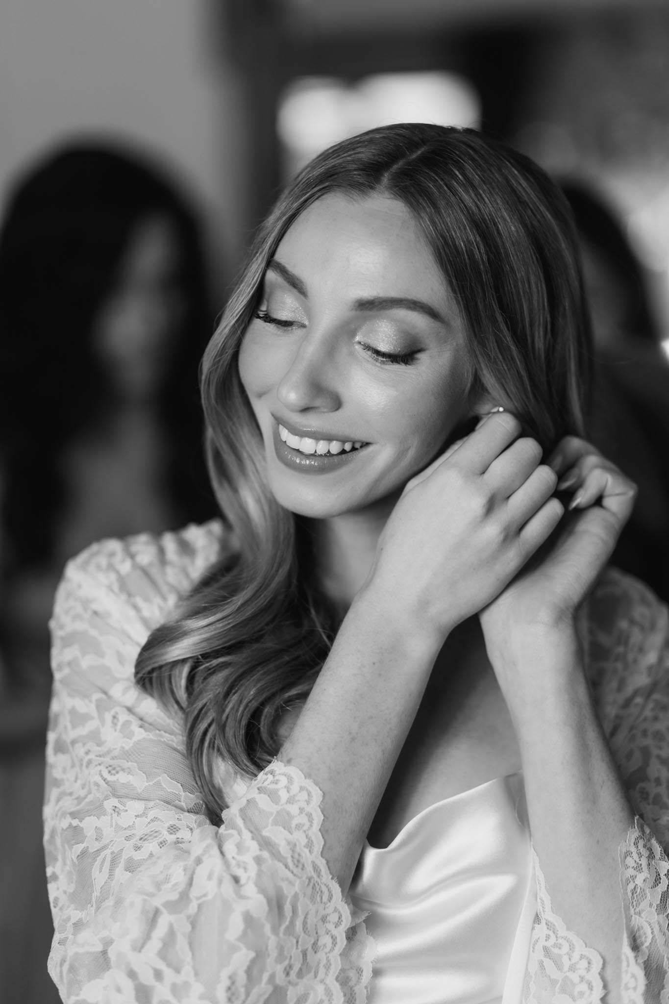 Black-and-white close-up portrait of a bride during the getting-ready phase, captured indoors with a softly blurred background showing at least one other person present. The bride is smiling with her eyes closed as she fastens an earring, her hands raised to her ear. She wears a satin bridal robe layered with a lace kimono-style cover-up with scalloped edges, and her hair is styled in loose waves. The image has high contrast with bright highlights on her skin and soft mid-tones throughout, giving it a polished, editorial feel.