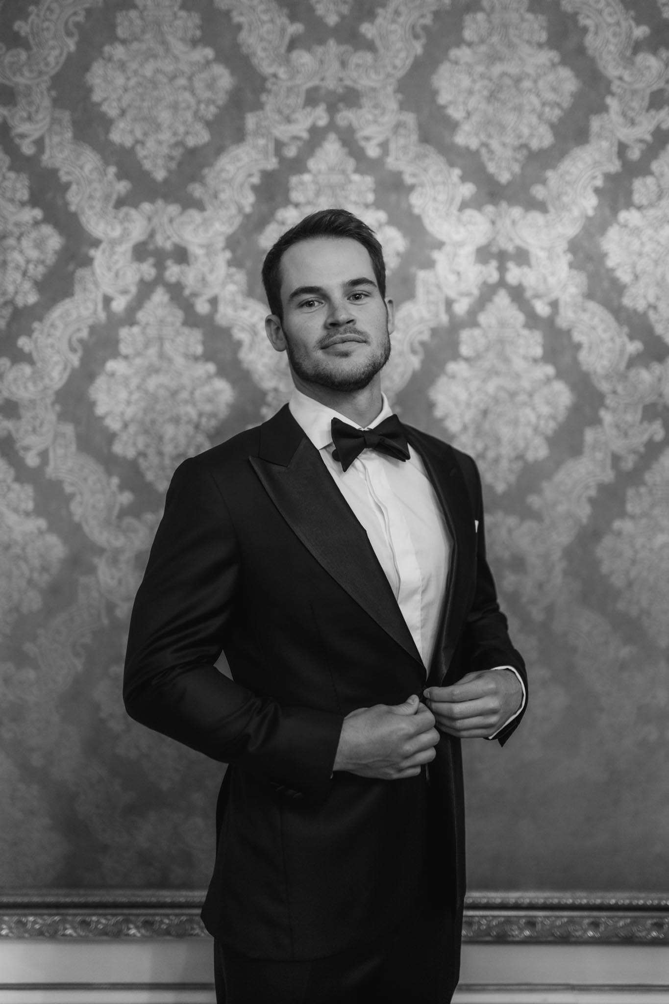 Groom adjusting tuxedo button in front of ornate damask wall with bow tie and pocket square, black and white