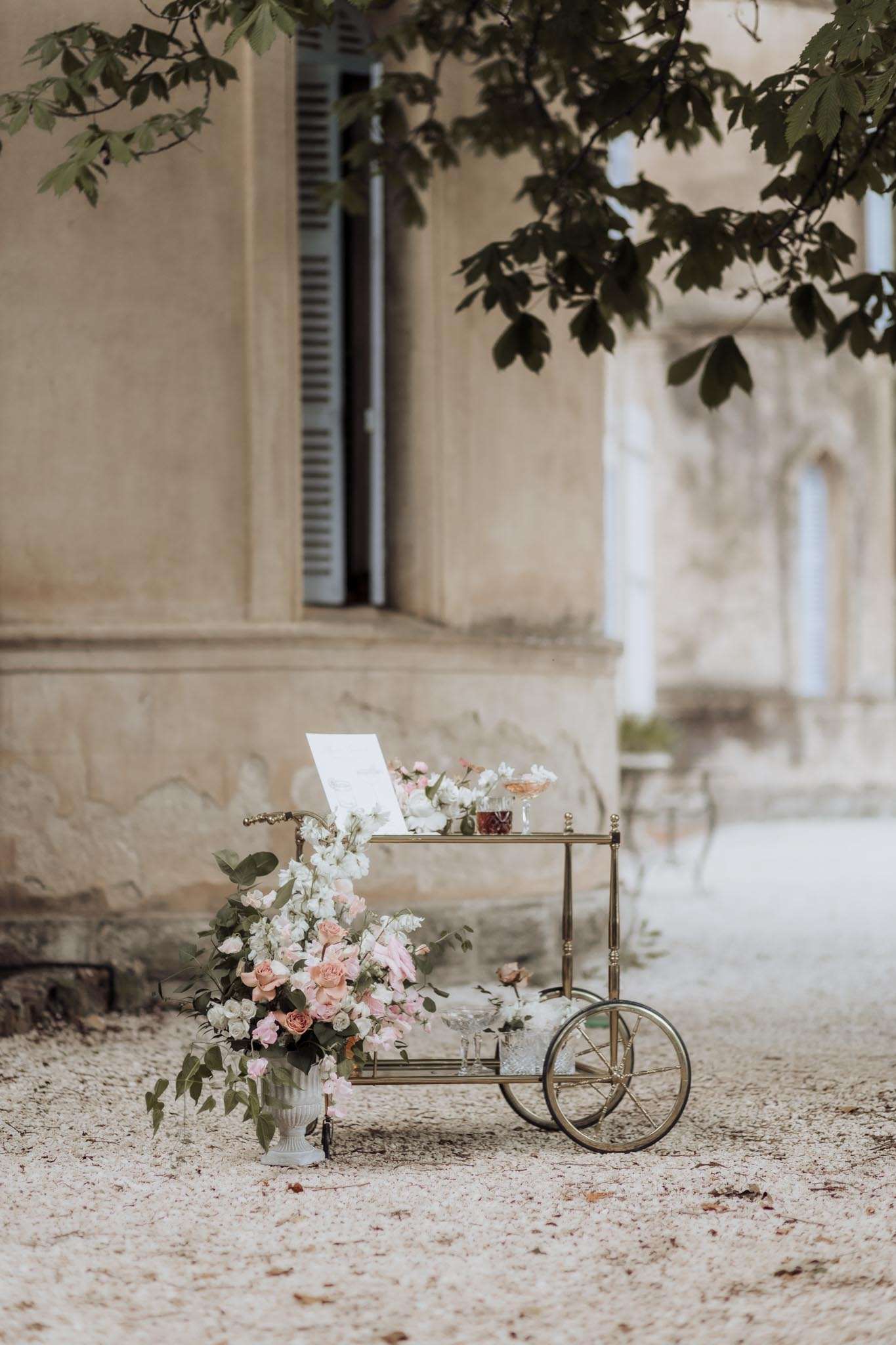 Brass bar cart with crystal coupes and menu card beside white urn of blush roses and trailing eucalyptus at chateau