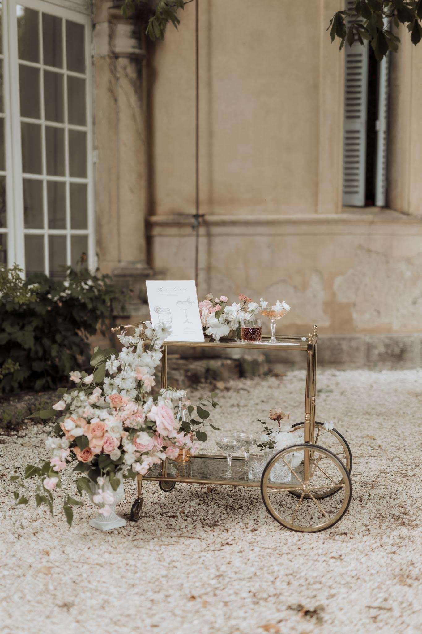 A styled bar cart setup photographed outdoors on a gravel courtyard in front of a classic French château facade with tall shuttered windows and stone columns. The two-tiered brass and glass bar cart is dressed with crystal glassware, amber-toned bottles, and cocktail glasses filled with blush-colored drinks, along with a printed cocktail menu card. White sweet peas and blush garden roses cascade over the top tier, while a large loose floral arrangement of blush and peach garden roses, white sweet peas, and eucalyptus spills from a white urn on the ground beside the cart. The lower tier holds additional crystal glasses and small bud vases. The overall decor palette is blush, peach, white, and gold with a classic French-romantic styling. Medium-distance detail shot with the château exterior softly out of focus in the background.