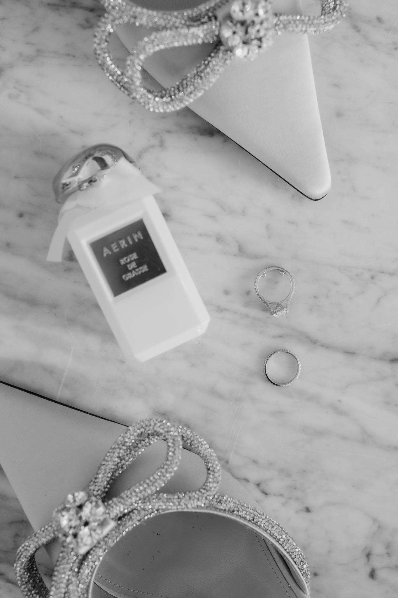 Black and white flat lay of crystal heeled sandals AERIN perfume and diamond rings on marble
