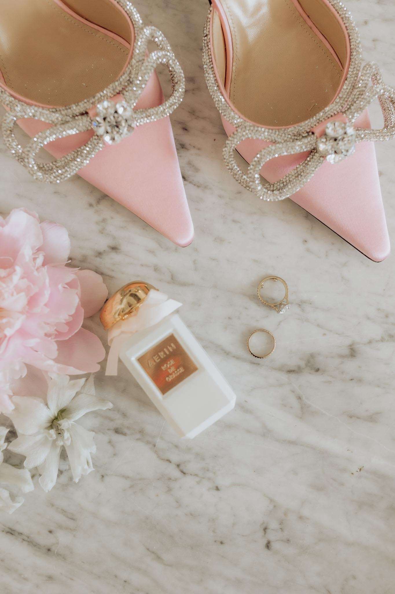 A bridal flat lay detail shot arranged on a white marble surface. The composition features a pair of blush pink pointed-toe heels with crystal-embellished straps and bow detailing at the toe, positioned at the top of the frame. Below the shoes are a small white and gold AERIN perfume bottle tied with a white ribbon bow, two gold rings — one diamond solitaire engagement ring and one slim gold band — and a loose blush pink peony alongside a white flower head, likely a ranunculus or small peony.