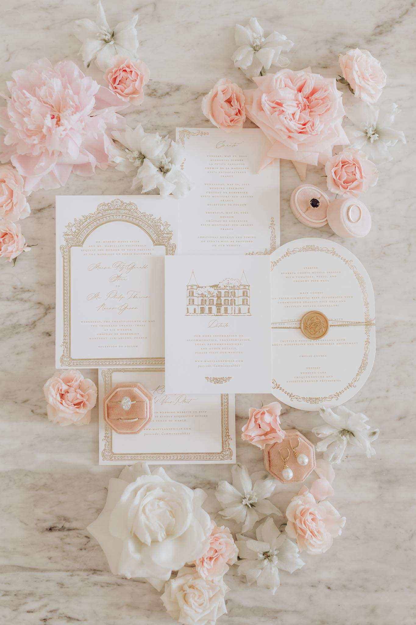 A flat lay detail shot of a complete wedding stationery suite arranged on a white marble surface. The suite includes a formal invitation with a gold ornate arch border, a details card featuring a line-drawn illustration of a French château, an events timeline card, a round menu card tied with a gold wax monogram seal, and a RSVP card — all printed in white with gold typography and decorative borders. Two blush velvet octagonal ring boxes are included in the arrangement, one holding a diamond solitaire engagement ring and the other containing pearl and gold drop earrings, with a third small blush ring box holding a dark stone ring nearby. The stationery suite is surrounded by loose blush garden roses, soft pink peonies, and white clematis blooms scattered across the marble. The overall palette is white, blush pink, and gold, consistent with a classic French château wedding aesthetic.