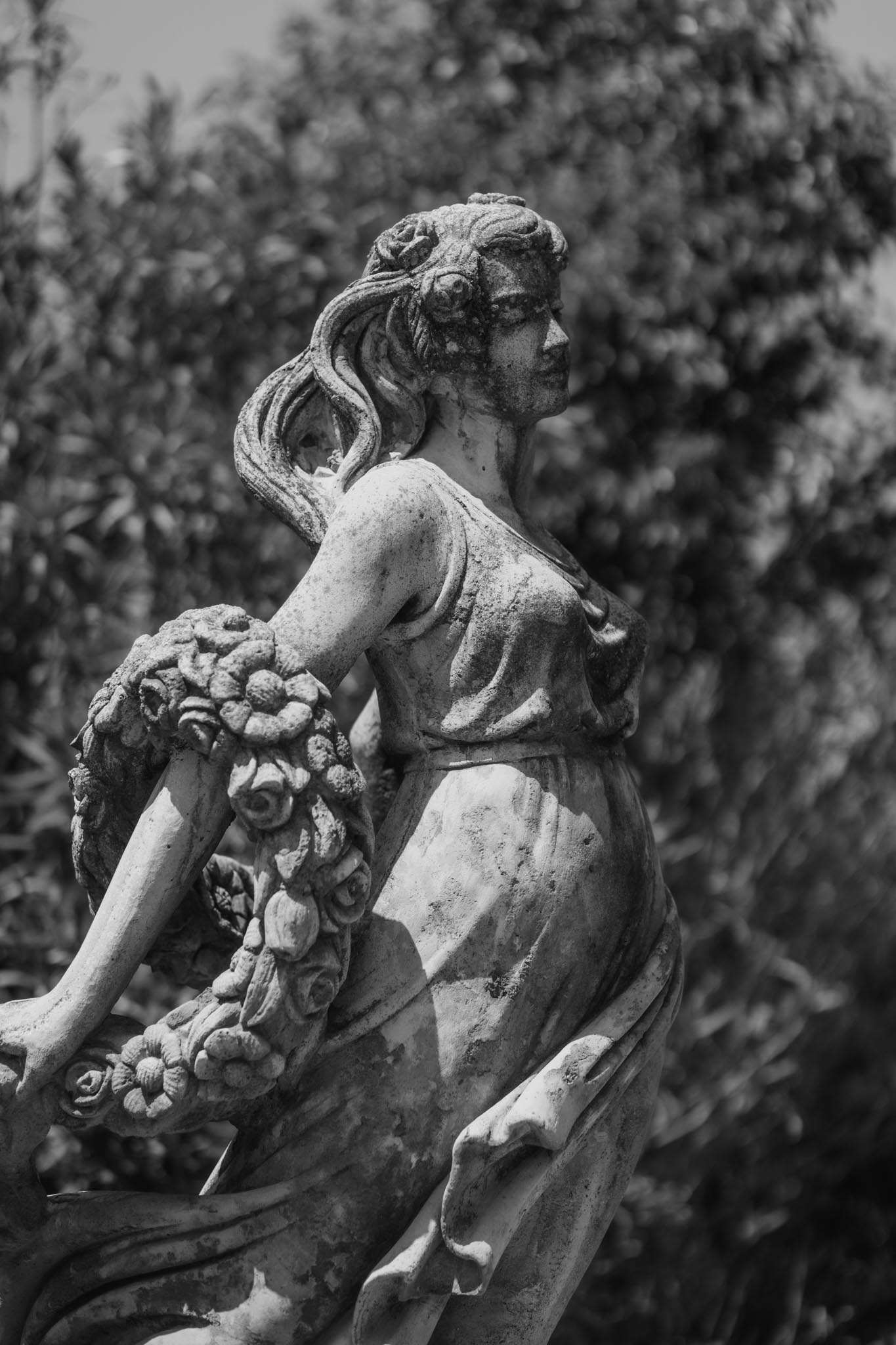 Black-and-white stone garden statue of female figure holding floral garland with roses and sunflowers in French garden